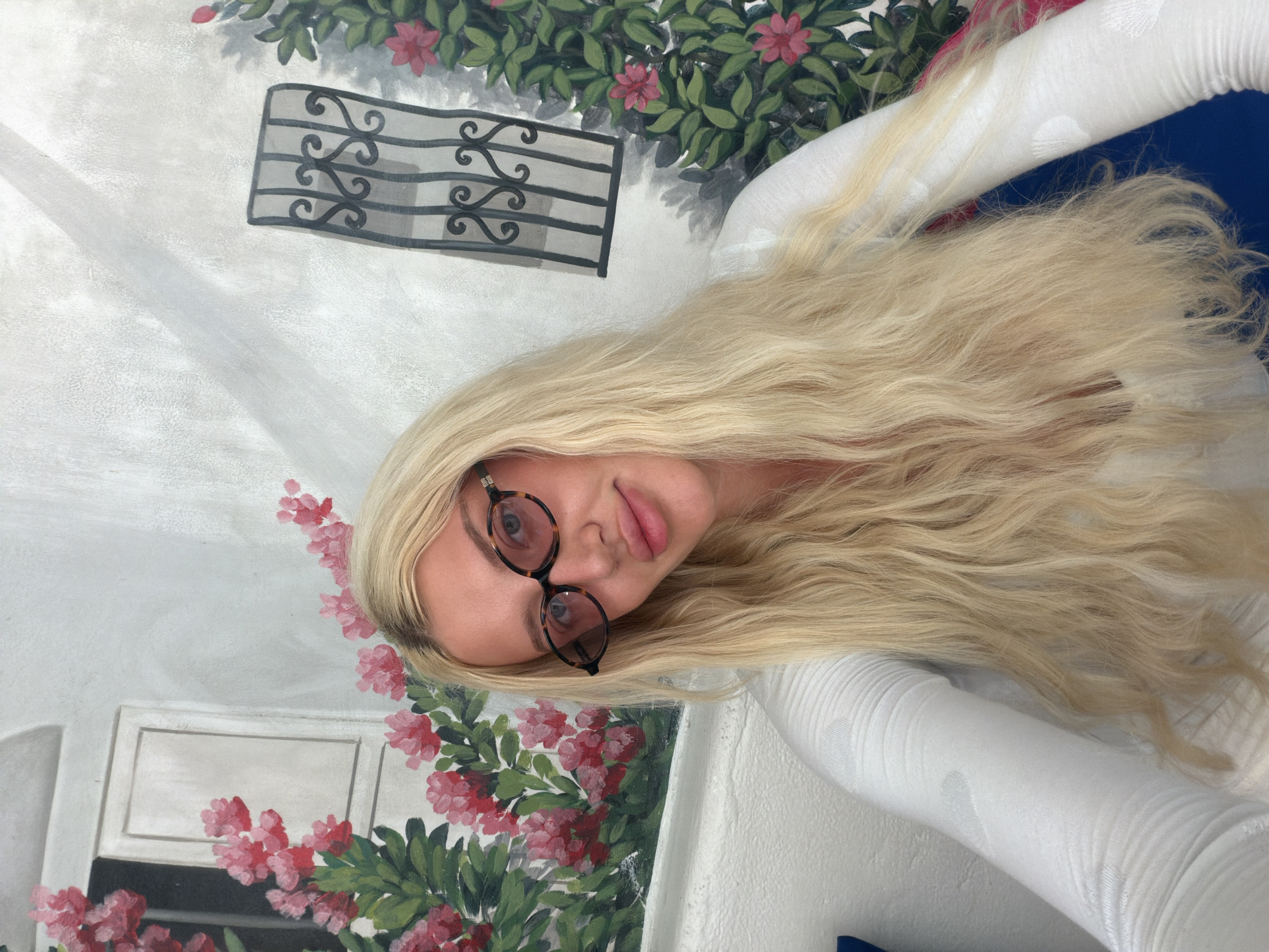 A young woman with long, blonde hair and tortoiseshell glasses is posing for a selfie against a mural. The mural depicts a white building with a window and red flowers on the left, and a black wrought-iron grate with greenery and pink flowers on the right. The woman is wearing a white, long-sleeved shirt. The setting appears to be outdoors, possibly in North Kuta, Indonesia, given the style of the mural which is reminiscent of Mediterranean architecture.