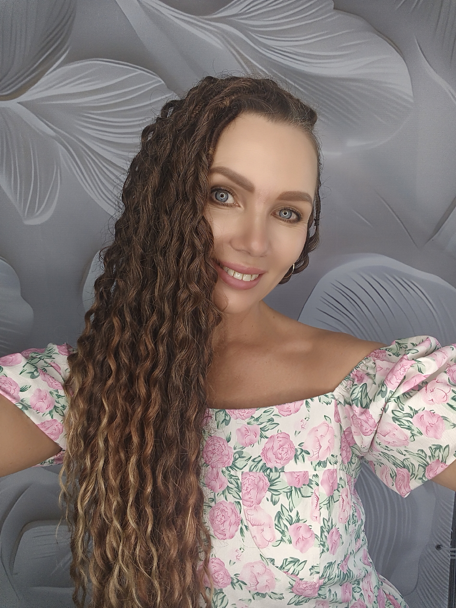 A woman is depicted in a selfie, her face angled slightly to the left, with her long, curly brown hair cascading down her left side. She has blue eyes and is smiling, revealing her teeth. She is wearing a floral-print dress with off-the-shoulder puff sleeves. The background is a textured grey wall with a floral pattern.