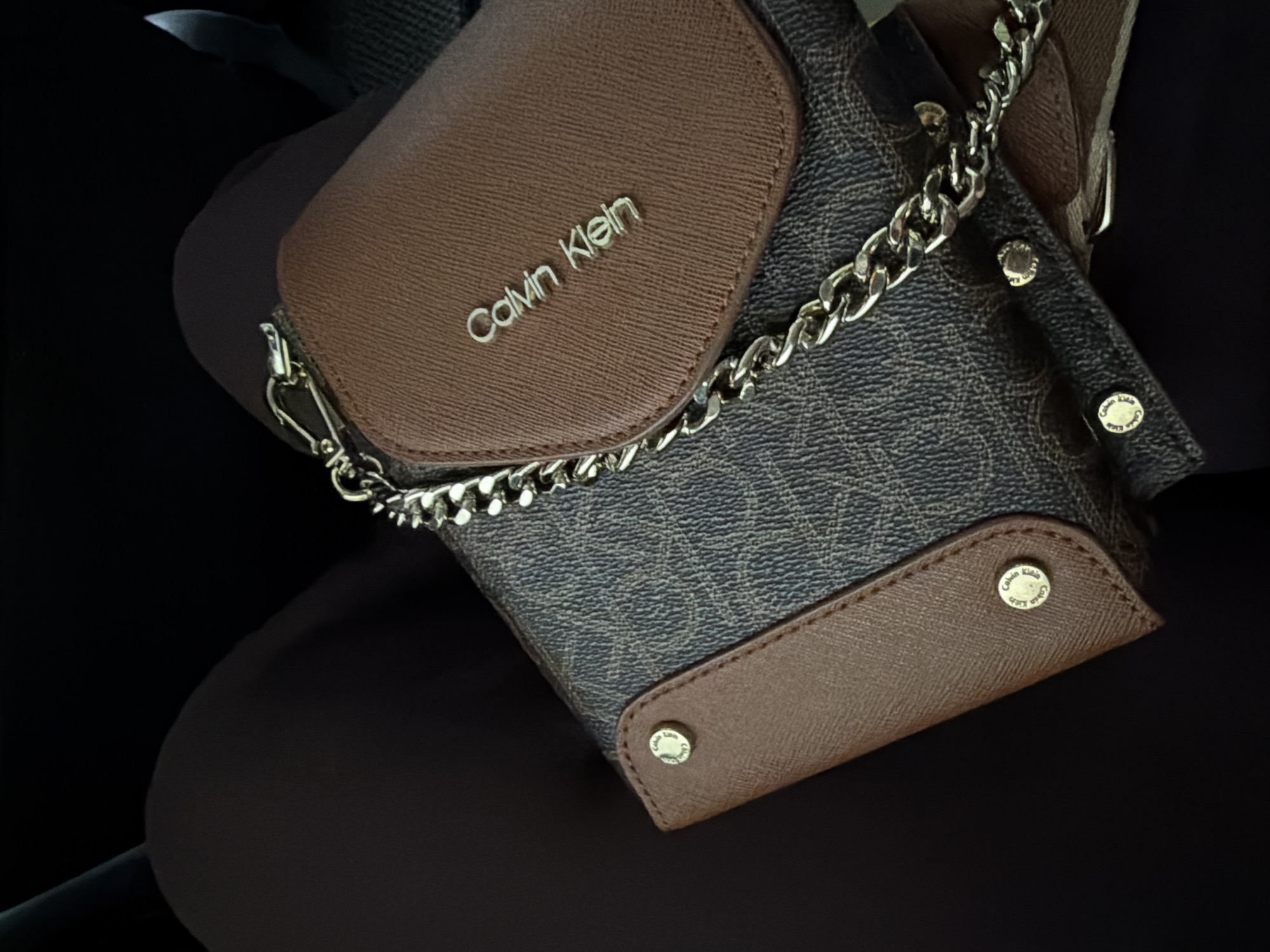 The image shows a Calvin Klein purse with a brown flap, a brown and black patterned body, and a silver chain. The purse appears to be resting on a dark surface. There is no information in the image to determine the location.