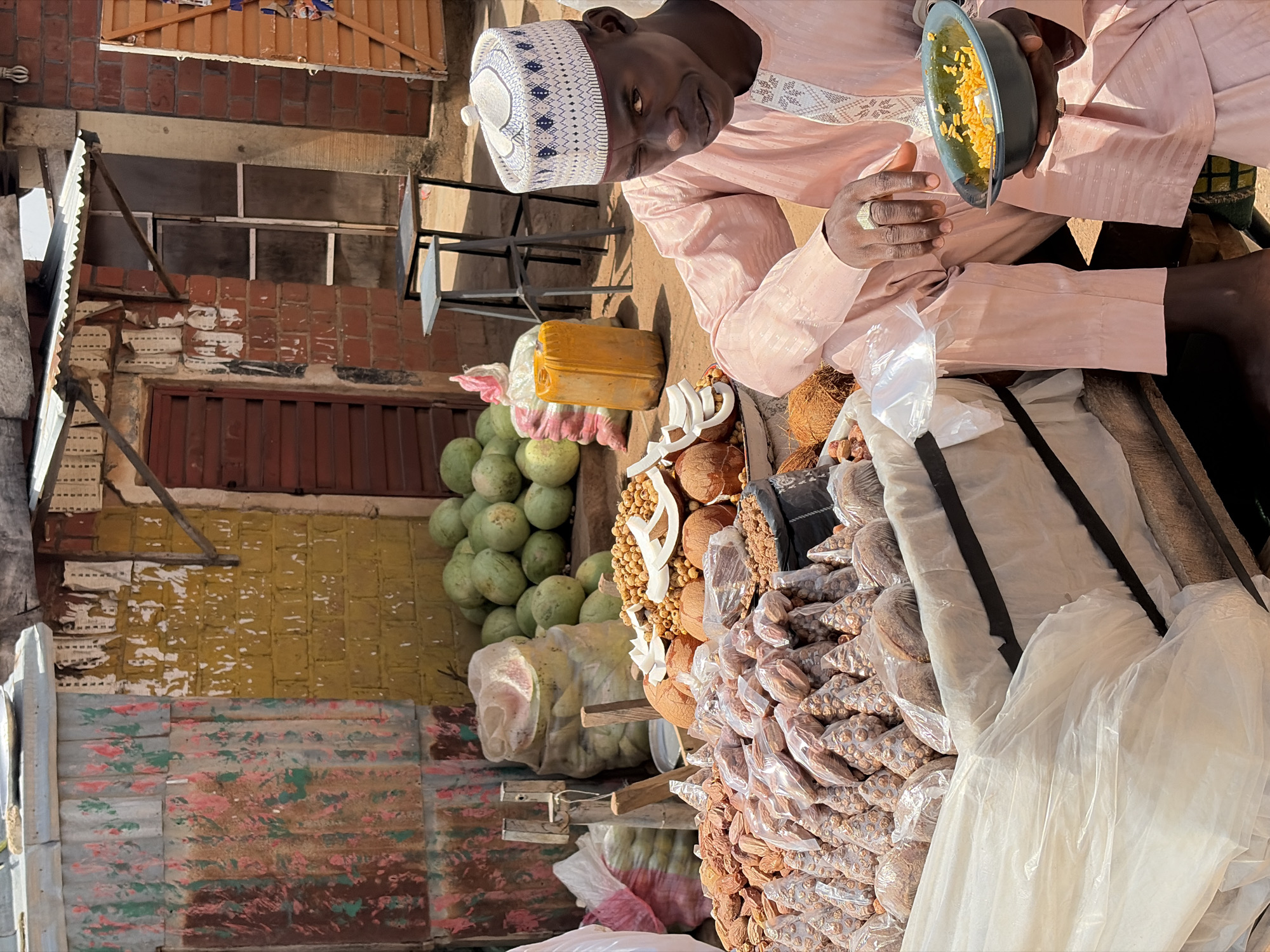 The image shows an overhead shot of a man in Gura Top, Nigeria, seated with a variety of goods displayed on a surface in front of him.
The man, wearing a pale pink outfit and a white and blue patterned kufi, holds a dark bowl with food inside. He has rings on his fingers.
In front of him, various items are laid out for sale, including a stack of round green fruits, possibly melons. Next to the fruits, there's a yellow container. There are several coconuts, some with white coconut meat exposed, along with other unidentifiable food items, many packaged in clear plastic bags. The goods are placed on top of a surface covered with a large white plastic sheet. The background shows a building with a mix of brick and yellow-painted walls, some metal frames, a closed wooden door, and a colorful patterned lower section.