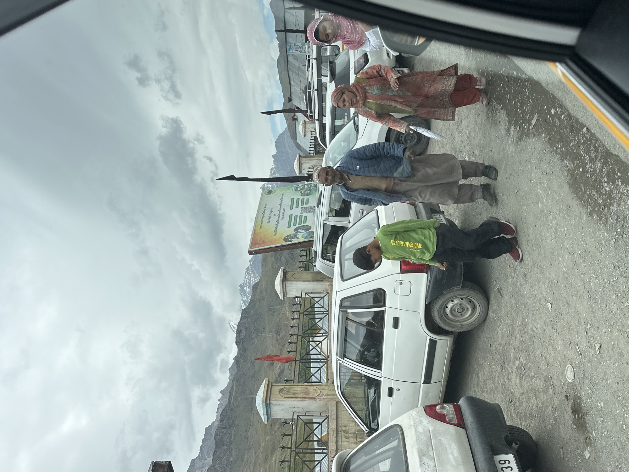 In Dras, India, under a cloudy sky, a group of people walk on a gravelly, wet road. The group consists of a young boy in a green long-sleeved shirt and dark pants, followed by an elderly man in a blue jacket and brown pants, and two women in colorful traditional clothing. One woman holds a white umbrella. Several cars are parked or stopped on the road, including a white Maruti 800. In the background, a fence with decorative pillars stands before a mountain range, with a red flag visible on a pole. A large banner with text and images is also present. The scene suggests a stop along a roadside in a mountainous region.