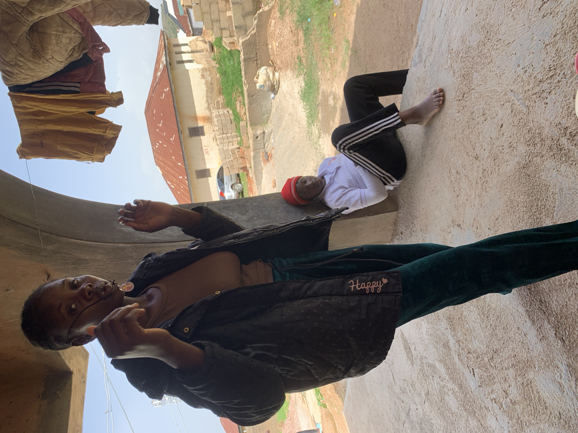 In Jos, Nigeria, a medium shot captures two individuals under an overhang. In the foreground, a woman stands and looks towards the camera, holding up a necklace. She wears a dark puffer jacket over a brown top and dark green velvet pants with "Happy" written on the leg. Behind her, a person sits on a concrete ledge, wearing a red beanie, white long-sleeved shirt, and black athletic pants with white stripes. The background reveals a rural or semi-urban scene with red-roofed buildings, construction materials, and laundry hanging on a line. The lighting suggests daytime.