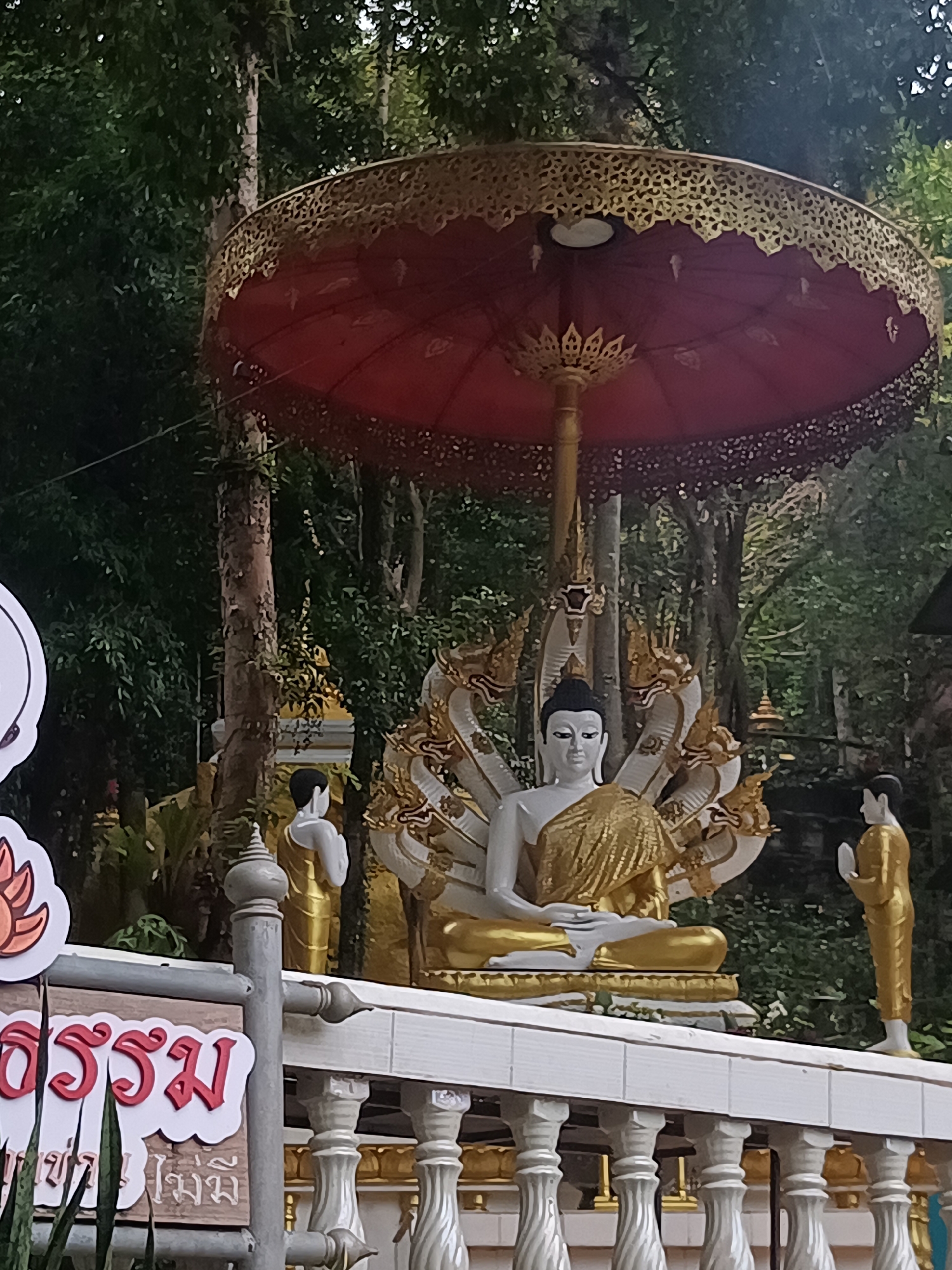 The image shows a statue of Buddha seated in meditation under a red and gold parasol, in ตำบลเกาะพะงัน, Thailand. The Buddha statue is primarily white with gold draping. Behind the Buddha is a backdrop of green foliage. Flanking the Buddha on either side are smaller, golden statues of figures in what appears to be a gesture of prayer. In the foreground, there is a sign in Thai script next to a white balustrade with gold trim. The setting suggests a temple or a sacred space.