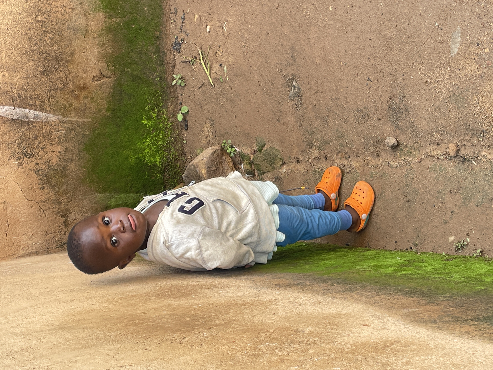A young Black child stands with their back to a weathered, light brown wall that is partially covered with patches of green moss. The child is wearing a light-colored hooded jacket with blue trim and a "GAP" logo on the front, a light blue undershirt, and blue pants. They are also wearing bright orange Crocs. The child is looking towards the camera with their tongue slightly sticking out. The ground is composed of dirt and small pebbles. In the background, to the right of the child, the ground slopes slightly upwards and there are some small green plants and debris. The overall environment appears to be outdoors in a simple, possibly rural or semi-urban setting, consistent with the area of Barakin Lerawi, Nigeria.