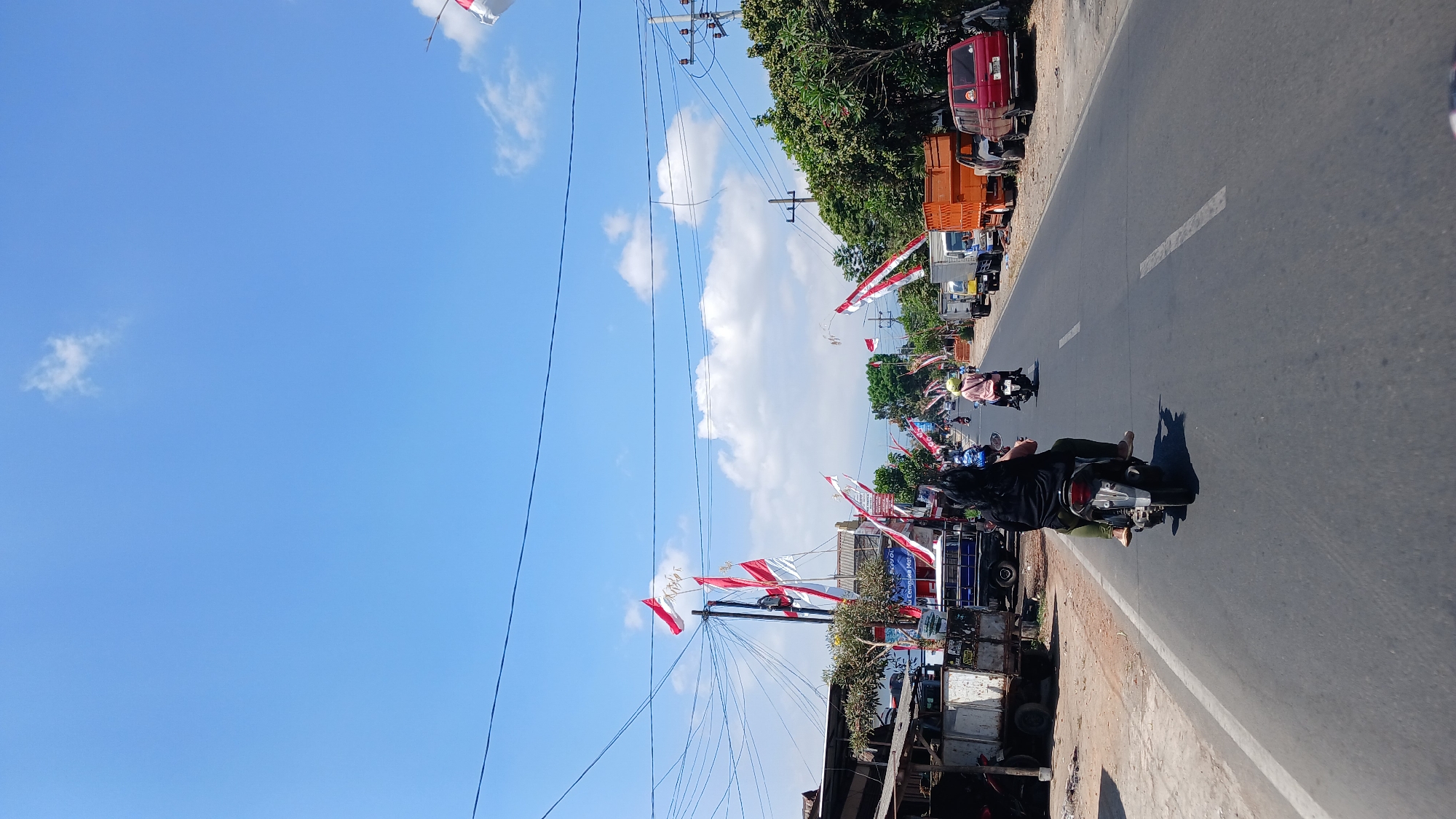 A daytime, vertical, eye-level, full shot shows a road in Pulungdowo, Indonesia, decorated with red and white Indonesian flags. The flags are strung across the road and mounted on poles along the side. The road is asphalt, with a white dashed line dividing the lanes. On the left side of the road, there are buildings and parked vehicles, including a white truck and a blue truck. On the right side of the road, there are trees, power poles, and more parked vehicles, including an orange truck and a red SUV. Several people on motorcycles are traveling on the road. In the foreground, a person wearing a black shirt and green pants is riding a motorcycle. The sky is blue with scattered white clouds.