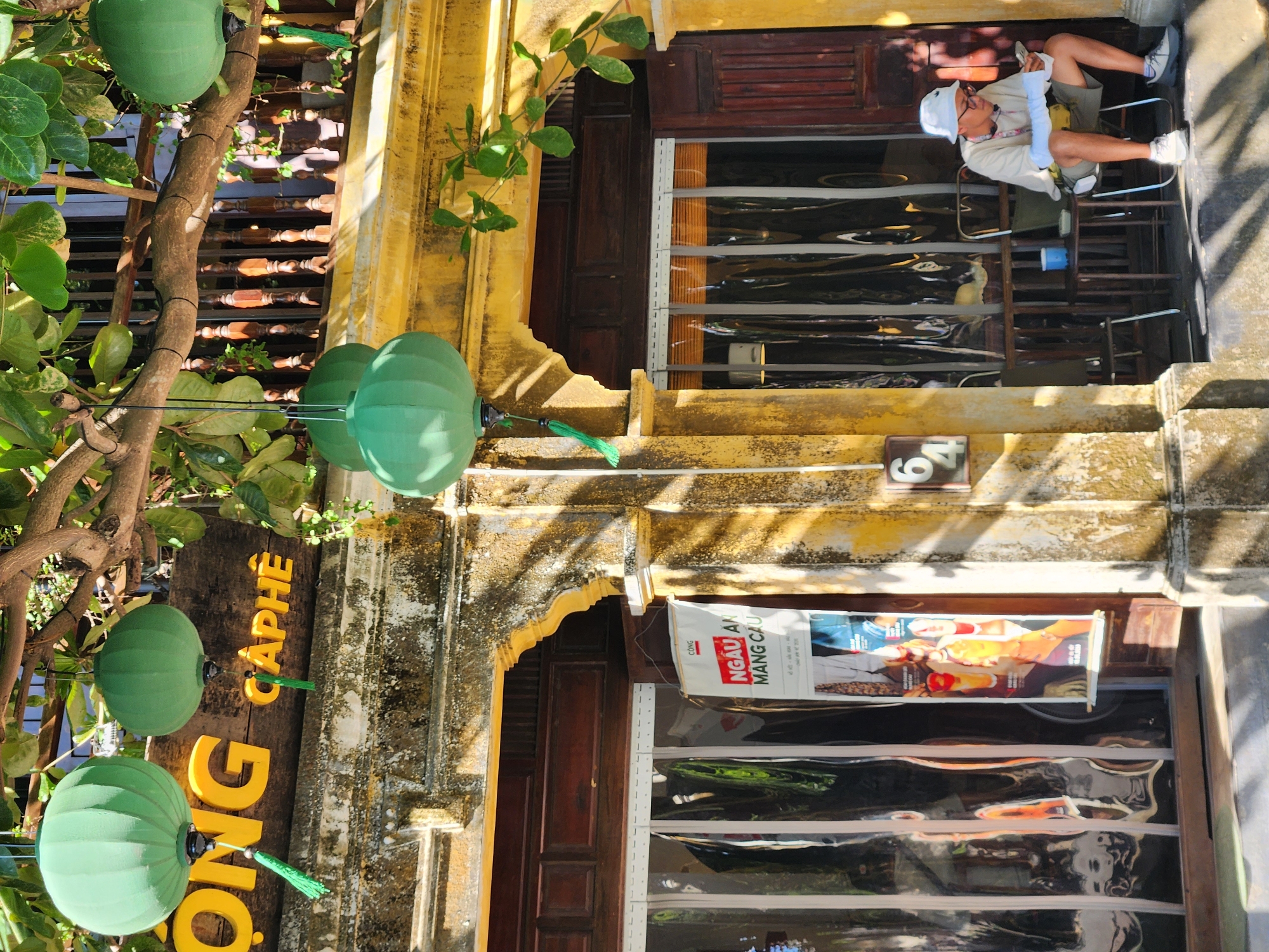 The image depicts a street scene in Hội An, Vietnam, bathed in bright sunlight, suggesting it's daytime. The overall impression is one of a warm, possibly tropical climate. The primary subject is a young person sitting on a chair in front of a building with yellow, weathered walls. The person is wearing a white hat and casual clothing, appearing relaxed and possibly observing the surroundings.
In the foreground, several large, green, spherical lanterns hang from a tree and are suspended by wires. These lanterns have green tassels and add a decorative element to the scene. A wooden sign with bright yellow lettering is partially visible, with the words "CAFÉ" and "NG" clearly legible. This suggests the presence of a cafe or restaurant.
The building itself has traditional wooden doors and shutters. A large window with a reflective surface is present, showing a distorted reflection of the interior or the opposite side of the street. A banner is hung on the building, featuring images of food and text, though the text is not entirely legible. A small sign with the number "64" is affixed to the wall near the entrance. The scene has a lively yet relaxed atmosphere, characteristic of many street scenes in Southeast Asia. The combination of the natural elements (tree, foliage) and the man-made structures (building, lanterns, sign) creates a visually rich and culturally specific setting.