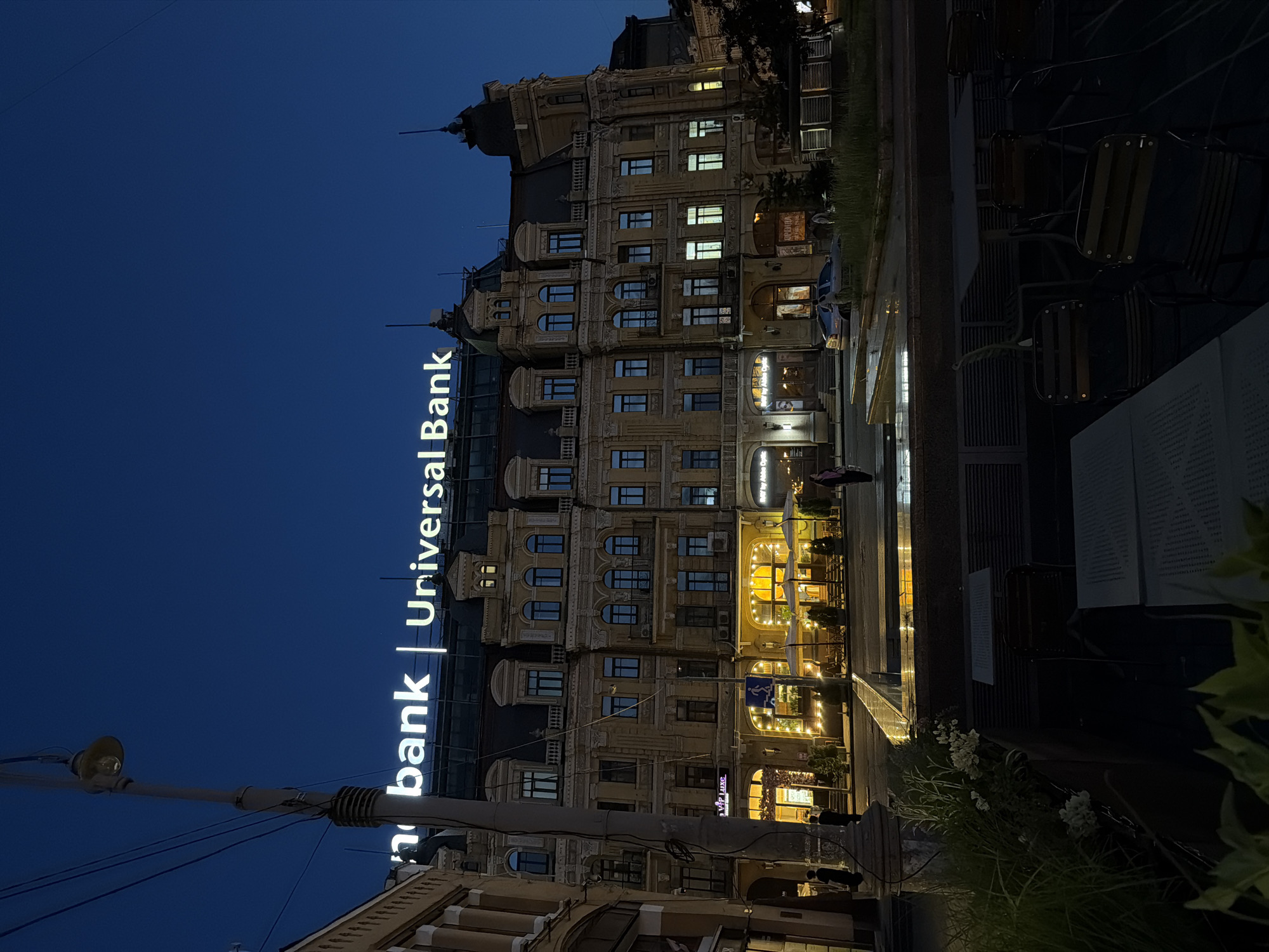 This is a nighttime photograph of a building in Kyiv, Ukraine. The building is lit up and has a sign that reads "Universal Bank". The street in front of the building is lined with tables and chairs, suggesting a restaurant or cafe. A few people are visible on the street, and there is a car parked nearby. The sky is dark blue, indicating that it is late evening or early morning. The architecture of the building is ornate, with detailed carvings and large windows.