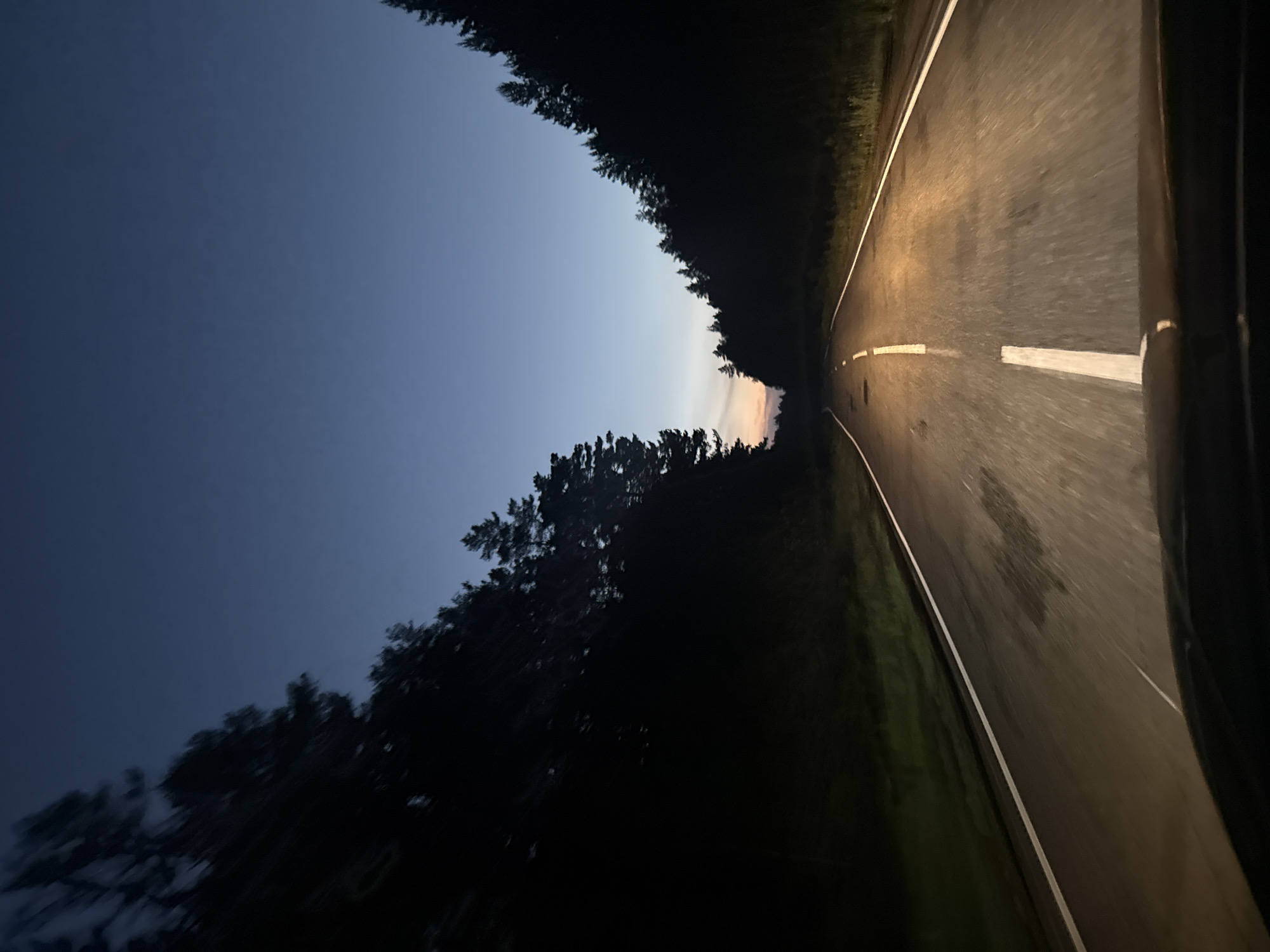 The image depicts a view from the perspective of a driver traveling on a two-lane asphalt road at dusk or night. The road has white lane markings. On either side of the road, there are dense, dark forests with tall coniferous trees. The sky above is a deep blue, transitioning to a lighter hue with a hint of orange near the horizon, suggesting the sun has recently set or is about to rise. The headlights of the vehicle illuminate the road ahead, casting a warm glow. The scene appears to be a rural or natural environment, likely a forested area. No people or specific landmarks indicating the city of Balak, Russia, are visible.