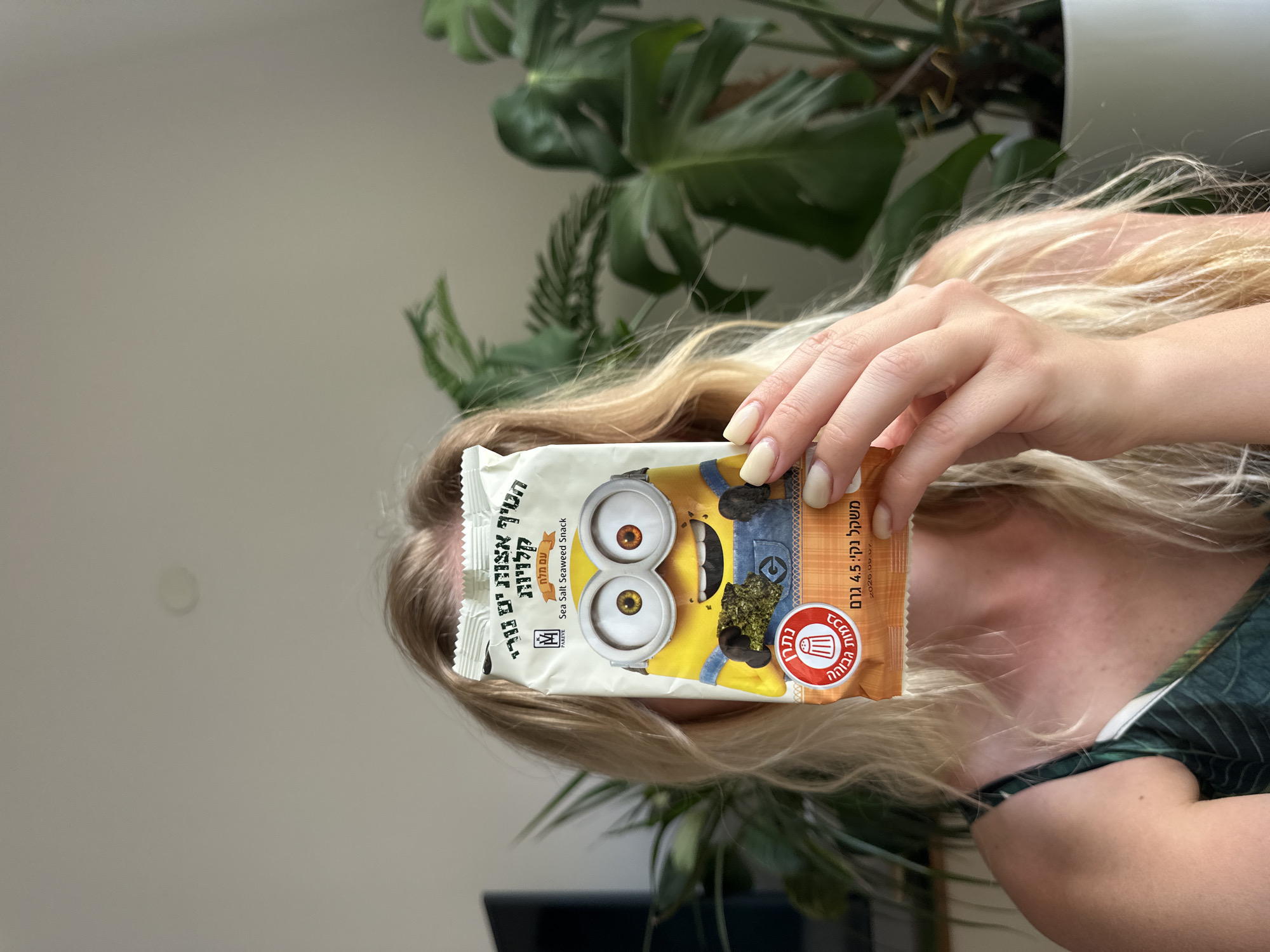 A blonde woman holds a snack bag featuring a Minion character in front of her face. The snack is Sea Salt Seaweed Snack, and the bag is primarily white with yellow and blue accents. The woman is wearing a green patterned top. She has light-colored fingernails. The background shows a potted plant with large green leaves, and a light-colored wall, consistent with an indoor residential setting, possibly in Petah Tikva, Israel.