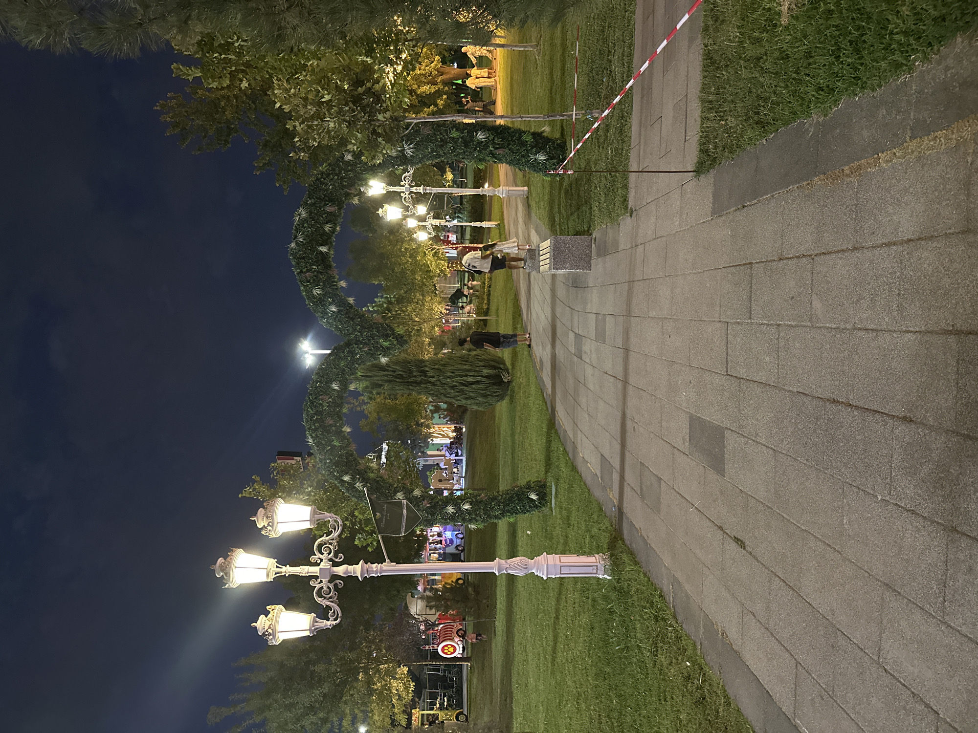 The image depicts a nighttime scene in a park, likely a public recreational area. The foreground is dominated by a paved walkway made of rectangular stone slabs, bordered by a strip of grass. To the left of the walkway, a tall, ornate lamppost with three glowing white lights stands prominently. Further into the park, two arched structures covered in greenery span across the walkway, illuminated by more lampposts and park lighting.
Several individuals are visible in the midground and background. On the walkway, a person wearing a black t-shirt and shorts stands to the left, facing away from the camera. A man and a young girl, both facing the camera, are standing near a stone bench in the middle ground, to the right of the walkway. Further back, in the right midground, a group of people, including a man in a white shirt and tan pants and another in a military-style uniform, are gathered. In the background, there are glimpses of what appear to be amusement park attractions, including a small train with brightly colored carriages.
The environment is characterized by well-maintained green lawns and mature trees. The sky is dark, indicating nighttime, and appears partly cloudy. A red and white striped tape, commonly used for crowd control or indicating a restricted area, is visible in the right midground, stretched between poles. The overall impression is of a lively park with families enjoying an evening out. This scene is consistent with the atmosphere of a park in Tashkent, Uzbekistan, known for its green spaces and family-friendly attractions, especially during evening hours when the weather is pleasant.