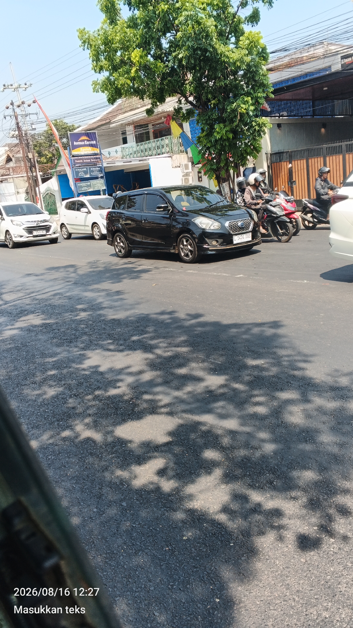 The image shows a street scene in Malang, Indonesia. A black car with license plate "L 1424 DBS" is in the foreground, stopped at an intersection. Behind it, two white cars are also stopped. Several motorcycles with riders are visible, one red and one black. In the background, there are buildings, trees, and power lines. The time stamp at the bottom left of the image indicates 2026/08/16 12:27. The text "Masukkan teks" is also present. The scene appears to be a typical urban street with traffic and pedestrians.
