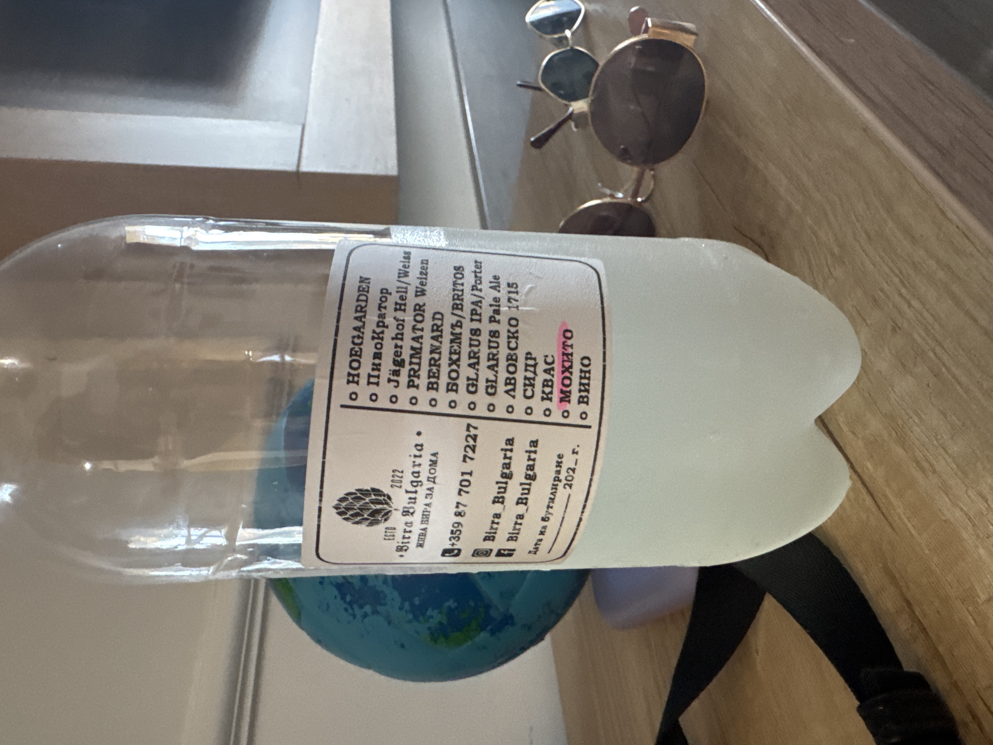 The image shows a clear plastic bottle filled with a pale liquid, likely beer or a similar beverage, standing on a wooden surface. A label is affixed to the bottle, featuring text in both English and Bulgarian, along with a graphic of hops. The label lists various beverage names, including "HOEGAARDEN," "ПивоКратор," "Jägerhof Hell/Weiss," "PRIMATOR Weizen," "BERNARD," "БОХЕМЬ/BRITOS," "GLARUS IPA/Porter," "GLARUS Pale Ale," "ЛВОЕCKO 1715," "СИДР," "КВАC," "МОХИТО," and "ВИНО." Below the list, there is contact information for "Birra Bulgaria" and a date of bottling "202_ г." To the left of the bottle, a portion of a blue globe is visible. In the background, a light-colored wall and a wooden surface are present. In the foreground, on the right side, a pair of sunglasses with gold frames and dark lenses are placed on the wooden surface. The image appears to have been taken indoors, likely on a shelf or table. The mention of "Birra Bulgaria" and the Bulgarian text on the label strongly suggest the location is within Bulgaria. The city of Nesebar is a historic town on the Black Sea coast of Bulgaria, known for its ancient ruins and modern tourism. While the image doesn't explicitly place the scene in Nesebar, the local context of a Bulgarian beverage producer makes it a plausible location for this photograph.