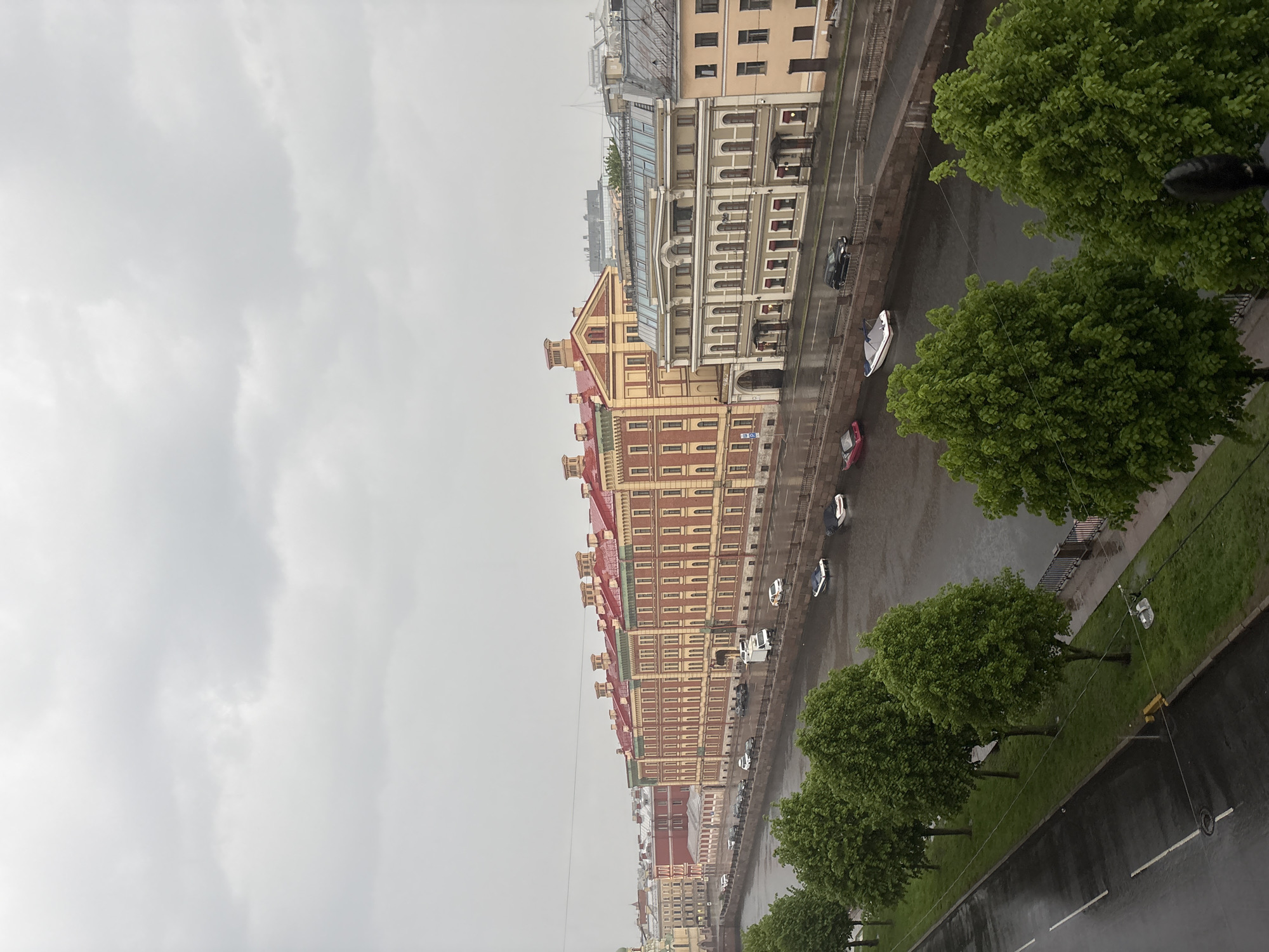 The image depicts a cityscape in Saint Petersburg, Russia, likely taken from an elevated position. The sky is overcast, suggesting rainy or cloudy weather. The view includes a canal lined with historic buildings, characterized by varying architectural styles and colors, predominantly earth tones and reds. Alongside the canal, there is a road with visible vehicular traffic, including cars and at least one boat. The foreground features trees lining the canal, their foliage appearing lush and green. The scene captures a typical urban environment in Saint Petersburg, blending its historical architecture with the natural elements of its canals and greenery.
