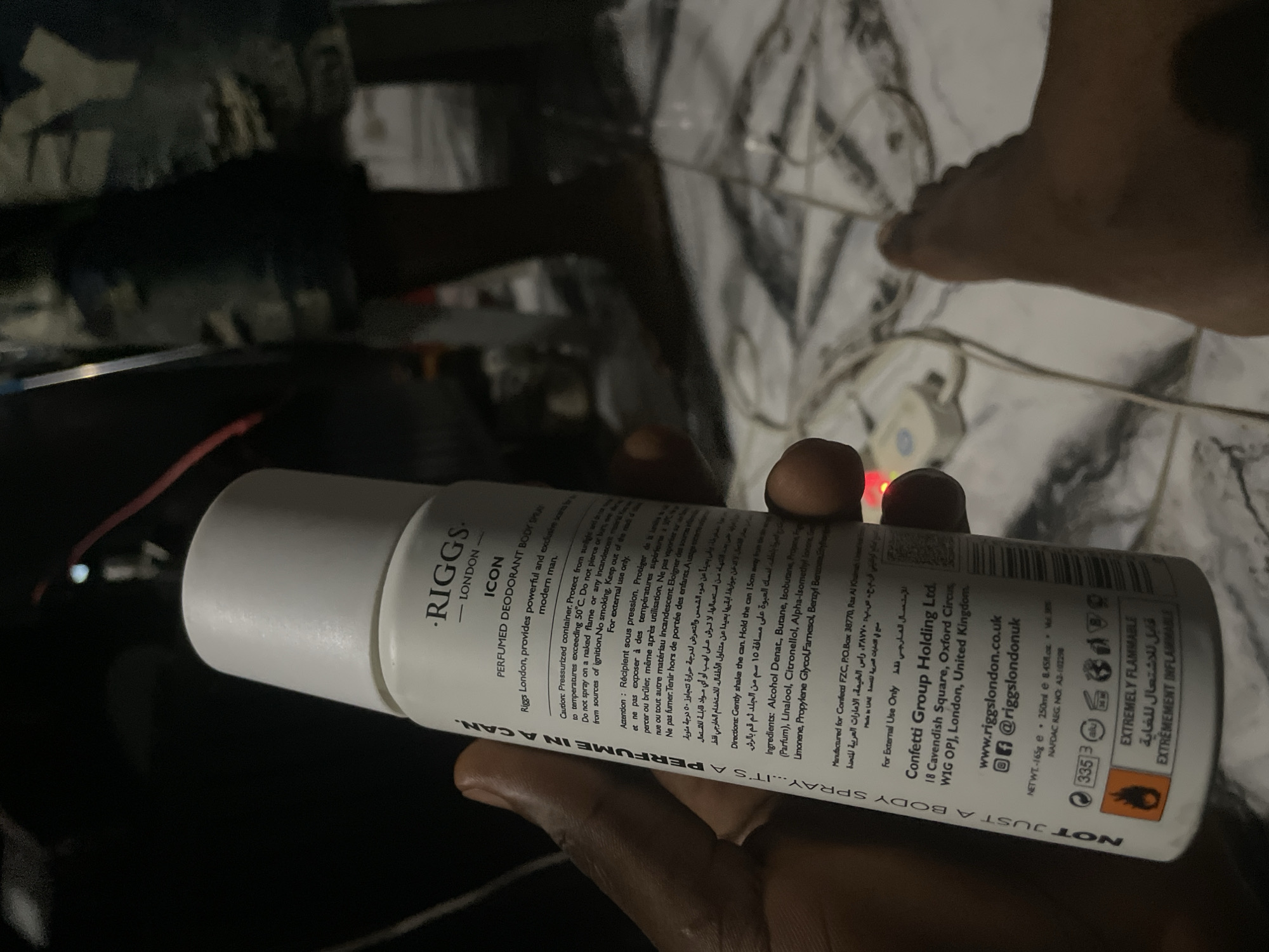The scene depicts a dimly lit indoor environment in Keffi, Nigeria. In the foreground, a dark-skinned human hand firmly grasps a white cylindrical aerosol can of "RIGGS - LONDON ICON PERFUMED DEODORANT BODY SPRAY." The can's label displays usage instructions, warnings, ingredients including Alcohol Denat and Butane, manufacturing details for Confetti FZC in Ras Al Khaimah, UAE, and Confetti Group Holding Ltd. in London, UK, and a NAFDAC REG. NO. A2-102298. A prominent "EXTREMELY FLAMMABLE" warning is visible with a flame pictogram.
The floor is a light-colored, patterned tiled surface, upon which numerous electrical wires, some coiled, are scattered. A small, light-colored electronic device, possibly a power control unit with a blue button, rests among the wires near the bottom center. In the lower right quadrant, a bare, dark-skinned human foot is partially visible, resting on the tiled floor. The background is dark and out of focus, with an indistinct blurred figure, possibly clad in camouflage-patterned attire, partially discernible in the upper right.