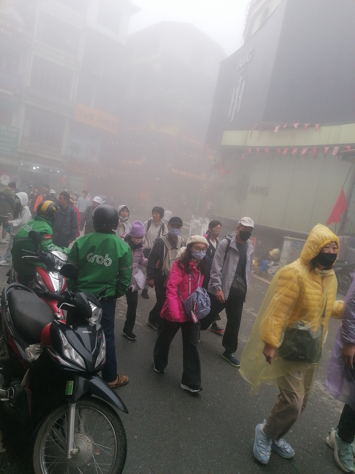 The image captures a bustling street scene in Sa Pa Ward, Vietnam, enveloped in dense fog or mist, significantly reducing visibility. Numerous pedestrians, many wearing face masks and warm clothing like jackets and hats, are walking on the wet asphalt road. One woman in the foreground wears a bright yellow plastic poncho, and another person on the right is partially visible in a purple poncho, suggesting rainy or damp conditions.
Two individuals in distinctive green jackets with the "Grab" logo are prominent on the left, one seated on a red and black Honda motorbike, and the other standing nearby. These figures indicate active ride-hailing or delivery services. Buildings line both sides of the street, appearing faded and indistinct due to the heavy fog. Visible text includes "AMG" on a large, dark building sign on the right, and "THE JADE" on a yellow sign on a building to the left. Red pennants or small flags are strung across the street or attached to buildings.
Despite the obscured environment, the scene appears active, with people moving purposefully. The time of day is likely daytime, given the diffused natural light. The overall mood is subdued due to the weather, but people are going about their activities, with some expressions appearing neutral, while the person in the yellow poncho shows a hint of a smile.