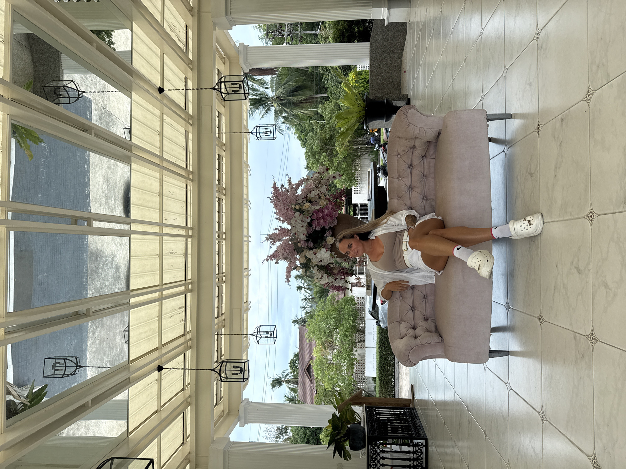 A woman is seated on a tufted pink sofa in what appears to be a covered outdoor entryway or lobby, with a view of lush tropical greenery and buildings in the background. The ceiling above is a reflective material, showing the sky and the underside of the structure. The woman is wearing a light brown tank top, white shorts, a white open cardigan, white socks, and white platform shoes. She is looking to her right with a slight smile. To her left, a large arrangement of pink and white flowers is visible. The floor is tiled in a light color with a repeating decorative pattern at the intersections of the tiles. The environment suggests a warm climate, consistent with Ko Samui District, Thailand, likely a resort or hotel entrance.