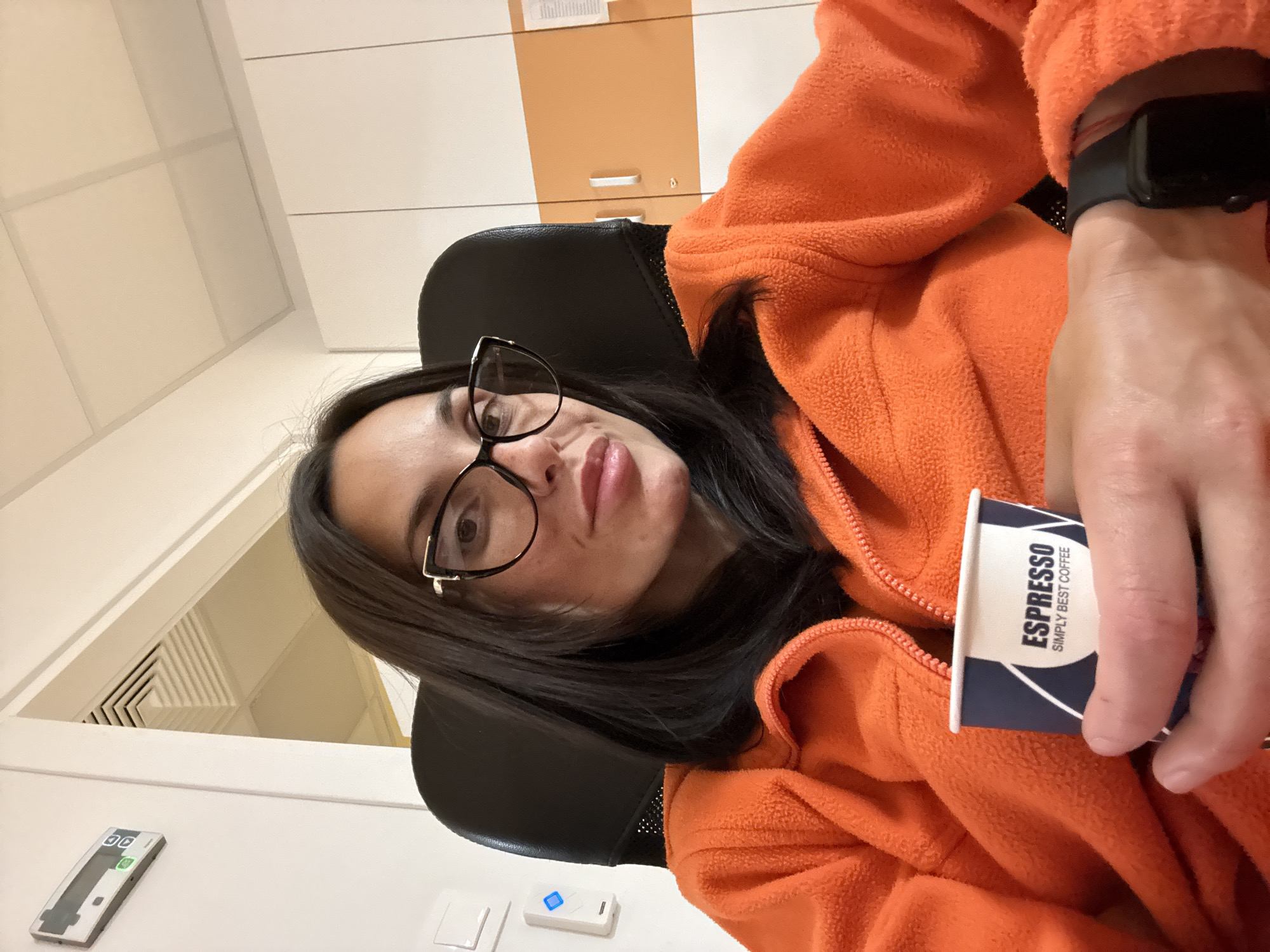 A woman with long, dark hair and glasses sits in a black office chair. She wears an orange fleece jacket. She holds a small cup of "Espresso Simply Best Coffee". She has a black smartwatch on her left wrist. The background shows white and orange cabinets, white walls with an air vent, and a white thermostat. There is no information to verify that the location is in the city of Pleven, Bulgaria.