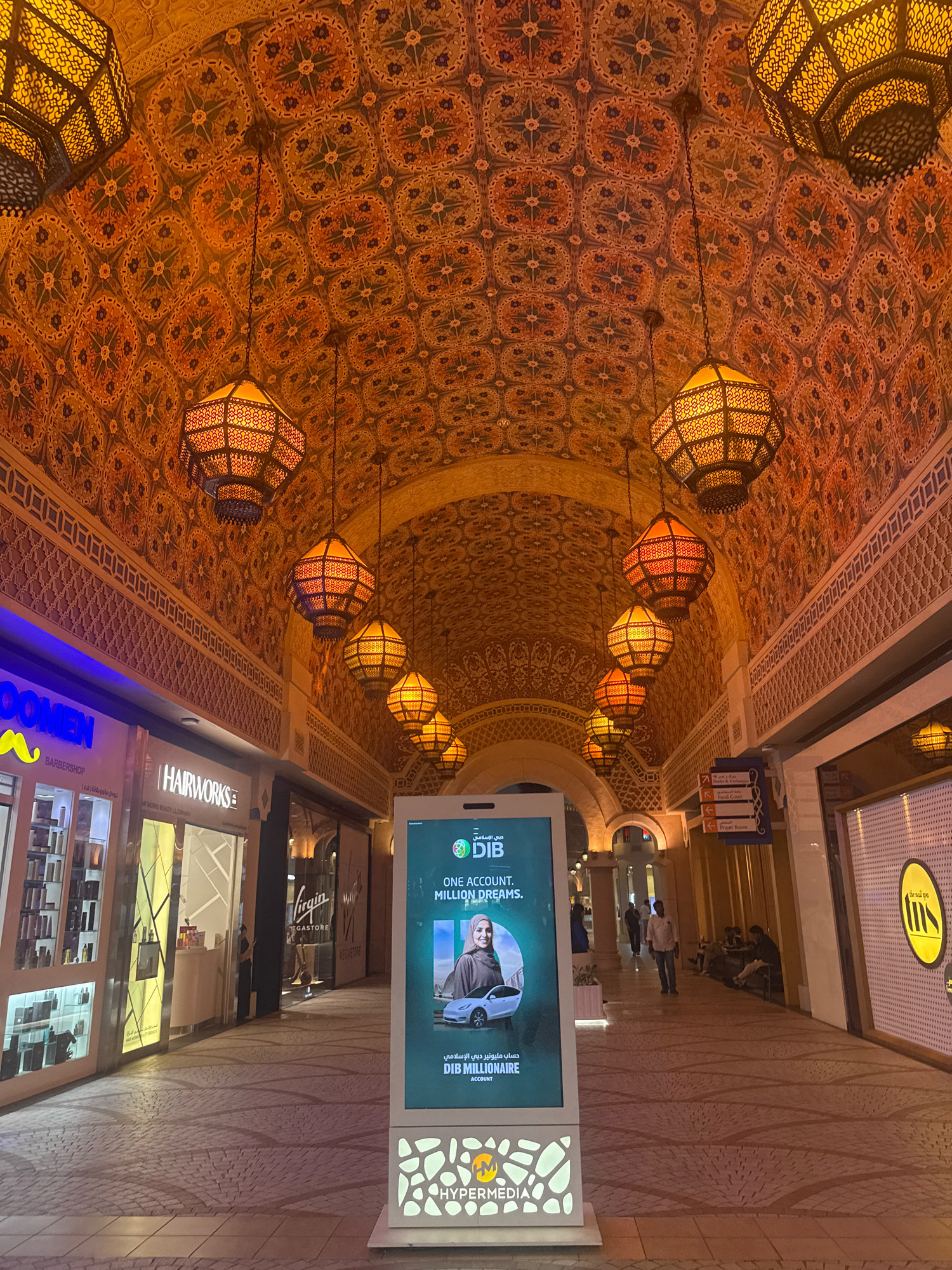 This image shows a well-lit indoor marketplace or shopping mall in Dubai, United Arab Emirates. The architecture features an ornate, patterned ceiling with a series of large, hexagonal, metal lanterns hanging from chains. The walls are decorated with intricate geometric patterns, and the shops lining the walkway have modern storefronts.
On the left, there is a shop with a blue and yellow sign that reads "OOMEN BARBERSHOP." Next to it is "HAIRWORKS." Further down the walkway, the "VIRGIN MEGASTORE" is visible.
In the center of the walkway, a large digital advertising screen displays a promotional campaign for "DIB MILLIONAIRE ACCOUNT" from DIB (Dubai Islamic Bank). The advertisement features a woman in a hijab next to a white car. Below the screen, the "HYPERMEDIA" logo is visible.
Several people are present in the scene. Some are walking down the corridor, while others are seated on benches or standing near the shops. The floor is paved with light-colored tiles arranged in a pattern. The overall atmosphere appears to be one of a busy commercial hub in Dubai, likely in a modern mall or a traditional souk with modern retail spaces.