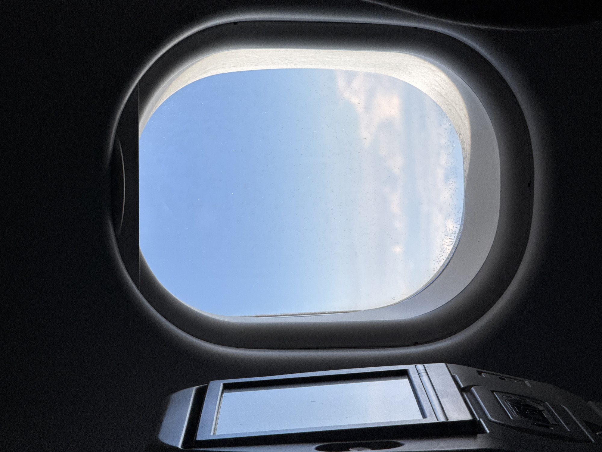 The image shows an interior view through a rounded window, likely from an aircraft or spacecraft. Through the window, a blue sky with light clouds is visible. Below the window is a dark-colored control panel or device with a small rectangular screen, reflecting the sky. The surrounding interior is dark and appears to be made of a smooth, possibly molded material. The location, Stolbishche, Russia, is not discernible from the image.