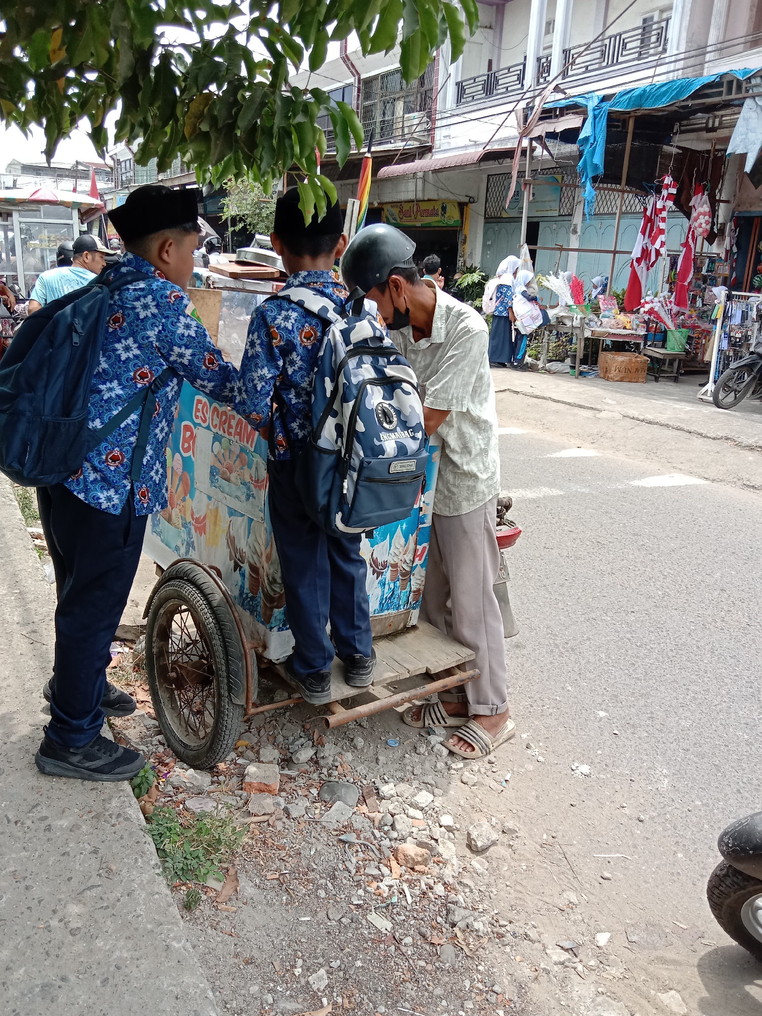 In Lhokseumawe, Indonesia, on a bright, sunny day, two young boys in matching blue patterned uniforms and black caps stand next to an ice cream cart. The cart's side displays a colorful advertisement featuring ice cream cones. One boy has a blue backpack with a white floral pattern, while the other sports a camouflage backpack. Behind them, a man in a light-colored patterned shirt and grey pants, wearing a black helmet, is leaning over the cart, possibly interacting with the boys or the vendor. The scene takes place on a paved street, with a sidewalk to the left littered with debris, including bricks and stones. Buildings with balconies line the street in the background, and stalls selling various goods, including flags and decorations, are visible. A motorcycle is parked on the right side of the image.