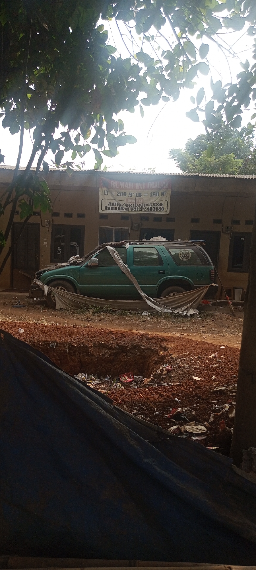 A green SUV is parked in front of a building. The SUV is partially covered with a light-colored tarp. A banner on the building reads "RUMAH INI DIJUAL" (This house is for sale) and provides property details: "LT = 200 M² LB = 180 M²" (Land area = 200 square meters, building area = 180 square meters), along with contact names "Anna" and "Ramadhan" and their phone numbers. The building appears to be a residential or commercial structure with a corrugated metal roof. In the foreground, a large dark tarp covers a pit filled with dirt and debris, suggesting ongoing construction or excavation. The ground around the SUV and building is unpaved and appears to be dirt and gravel. The scene is outdoors, with trees and foliage visible in the background, under a bright, hazy sky. The overall impression is one of a property being advertised for sale, possibly in a state of disrepair or under development.