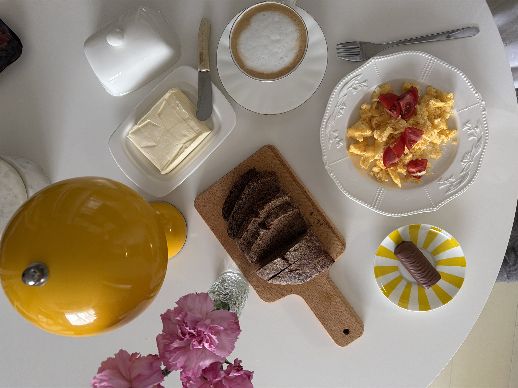 This overhead shot captures a table set for a meal, likely breakfast or brunch, in Khimki, Russia. The white table surface is laden with various food items and tableware, suggesting a domestic setting.
In the upper right, a white ceramic plate with a decorative rim holds a portion of scrambled eggs, garnished with chopped red tomatoes and a sprinkle of a light-colored topping, possibly cheese. A silver fork rests above the plate. To the left of the eggs, a white cup filled with coffee topped with thick white foam sits on a matching saucer.
A white ceramic butter dish contains a block of butter, showing impressions from a knife. A small knife with a light wooden handle is placed next to the butter. The lid for the butter dish is situated near the top left corner of the frame.
Centrally positioned, a light wooden cutting board displays several slices of dark, rustic bread. Below the bread and to the left, a large, glossy yellow spherical object, possibly a kettle or a pot, is visible. Adjacent to it, a small glass vase holds vibrant pink carnation flowers.
In the lower right quadrant, a small round plate with a distinctive yellow and white radial stripe pattern holds a single, dark brown chocolate bar with a ribbed texture.
No people are present in the image, and no visible text can be deciphered. The arrangement suggests an inviting and colorful spread for a morning or early afternoon meal.