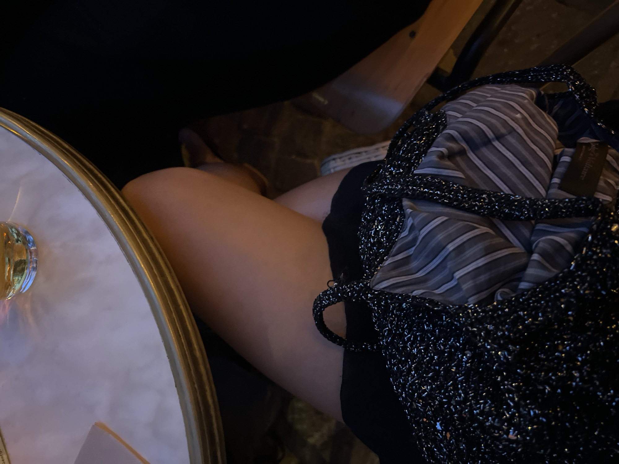 The image shows a close-up view of a person's legs and a handbag, positioned at a table in an outdoor Parisian cafe. The table has a light-colored, marbled surface with a gold-colored rim. A glass of amber liquid, likely a beverage, is visible on the table. The handbag, made of a dark, sparkly material, has a striped lining. The person is wearing a black outfit and a light-colored shoe is partially visible. The background consists of dark shadows and a wooden chair. The scene suggests a relaxed setting, possibly a social gathering or a moment of personal reflection. The presence of a glass of drink and the cafe ambiance places the scene in Paris, France.
