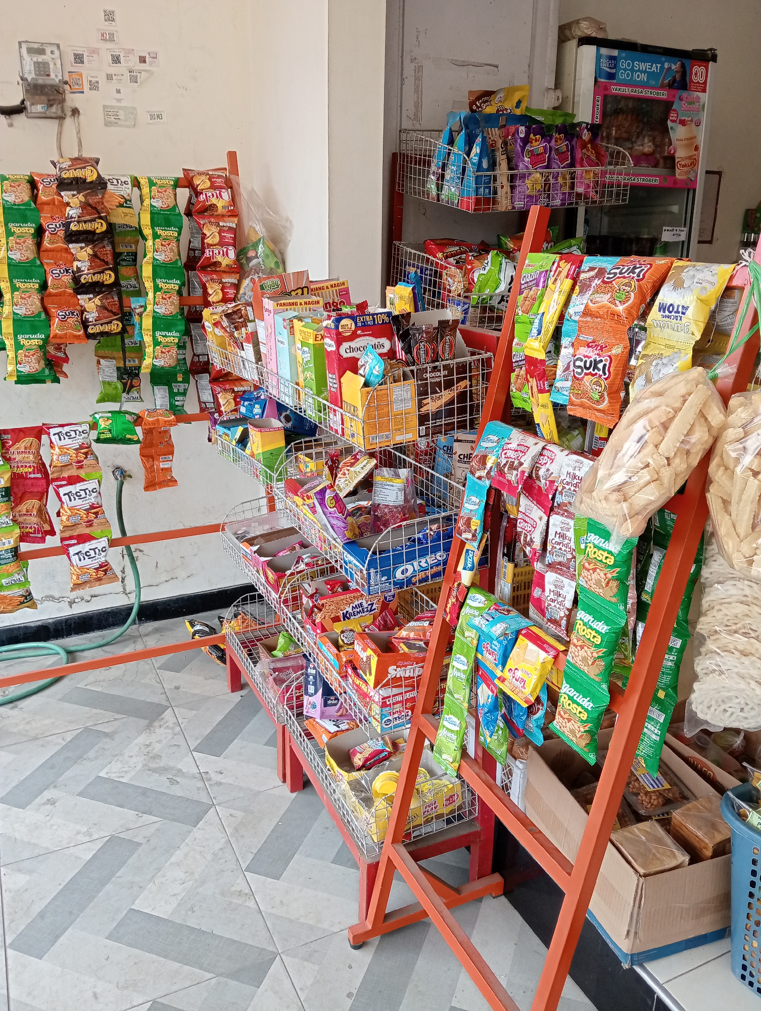 The image depicts the interior of a small convenience store or warung, likely located in Grabag, Indonesia, based on contextual cues. The store is filled with a variety of packaged snacks and beverages displayed on metal shelving units and racks.
In the foreground, an orange metal display rack holds multiple tiers of bagged snacks. These include brands such as "Garuda Rosta," "TicTac," and various colorful packets of chips and crackers. Some snacks are hung on hooks, while others are placed in wire baskets. To the right, a light blue plastic basket contains items, and several cardboard boxes are visible on the floor, some containing packaged goods.
The background shows a white wall with a vintage electricity meter mounted on it, along with several small printed QR codes. A green garden hose is draped on the floor near the wall. Further into the store, a refrigerator unit is visible with advertisements for "GO SWEAT GO ION" and "Yakult Rasa Stroberi." The floor is tiled with a geometric pattern of grey and off-white tiles. The overall scene suggests a typical small retail environment focused on selling fast-moving consumer goods.