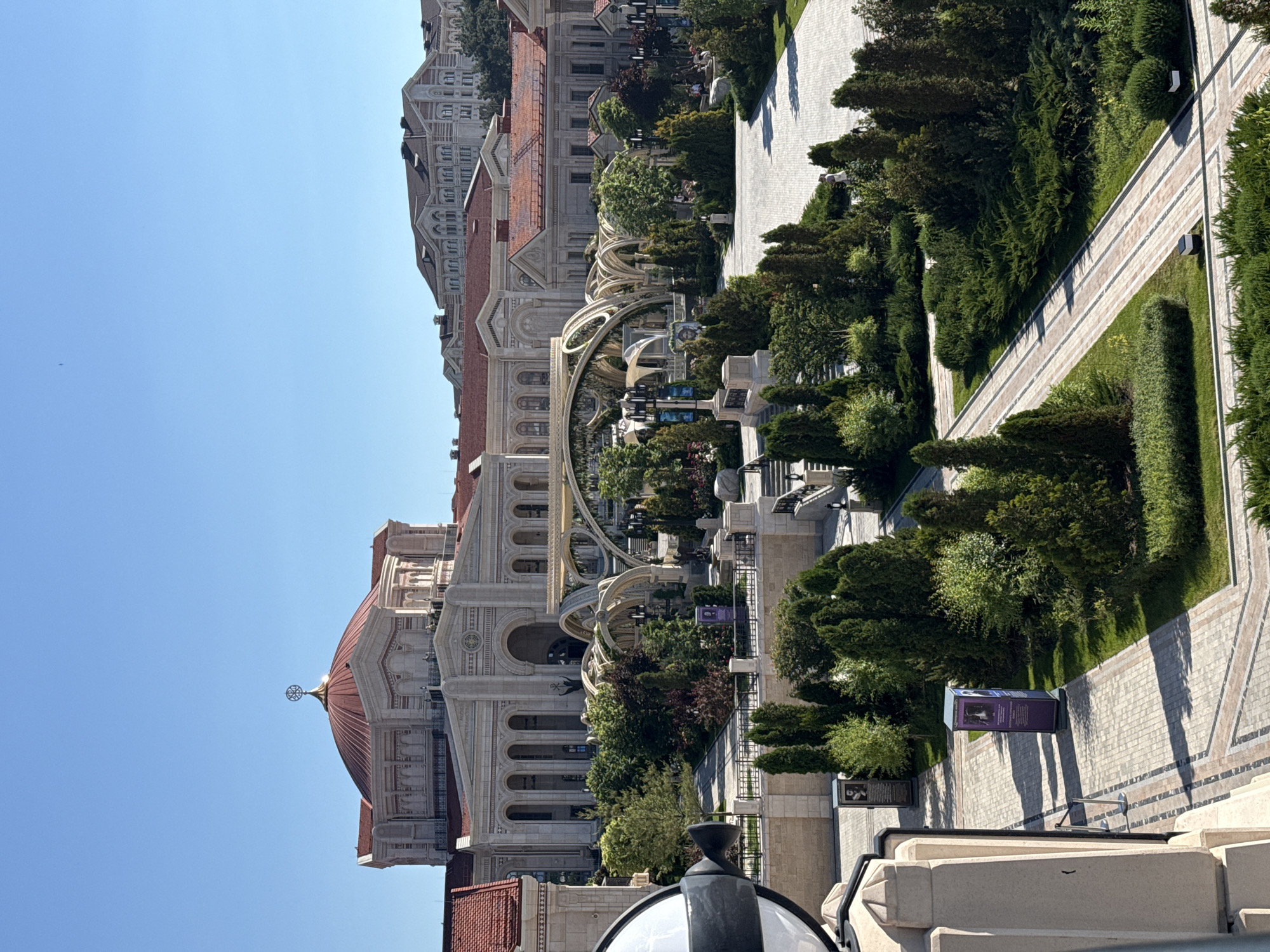 The image shows a sunny day with a clear blue sky over a large, ornate building with a red dome and columns. In front of the building is a landscaped park area with trees, bushes, and paved walkways. There are also some modern-looking architectural structures with arched elements. Several people are visible in the park, walking and standing. The overall scene depicts an outdoor public space, likely a tourist attraction or a cultural center.