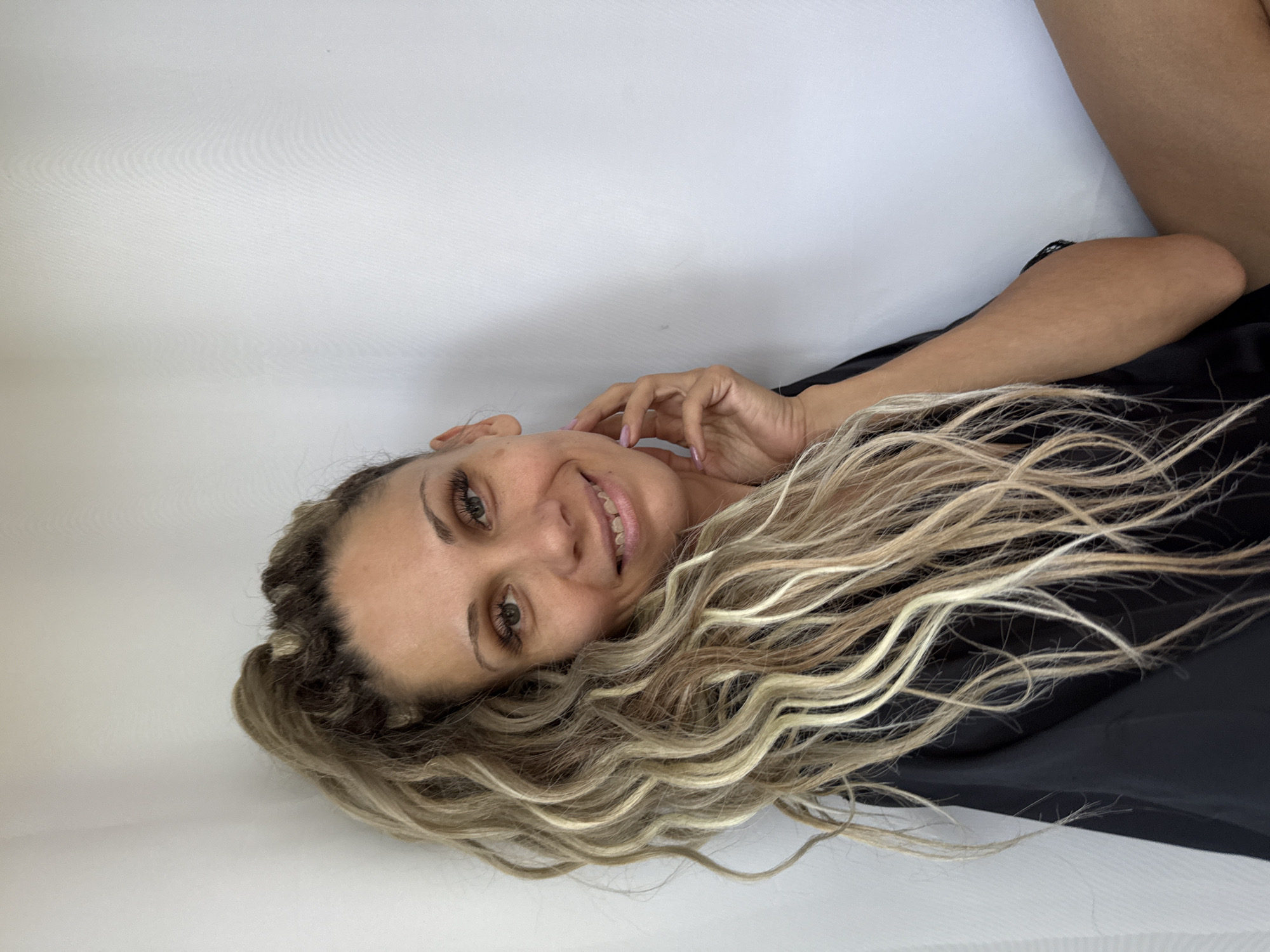 A blonde woman with long, curly hair tied into a high ponytail is leaning against a white wall. She is wearing a black robe. Her right hand is touching her face. She is smiling. The image appears to have been taken indoors, possibly in a studio setting given the plain white background. There is no indication of the specific location within Rusanovo, Russia, or any other details of the environment.
