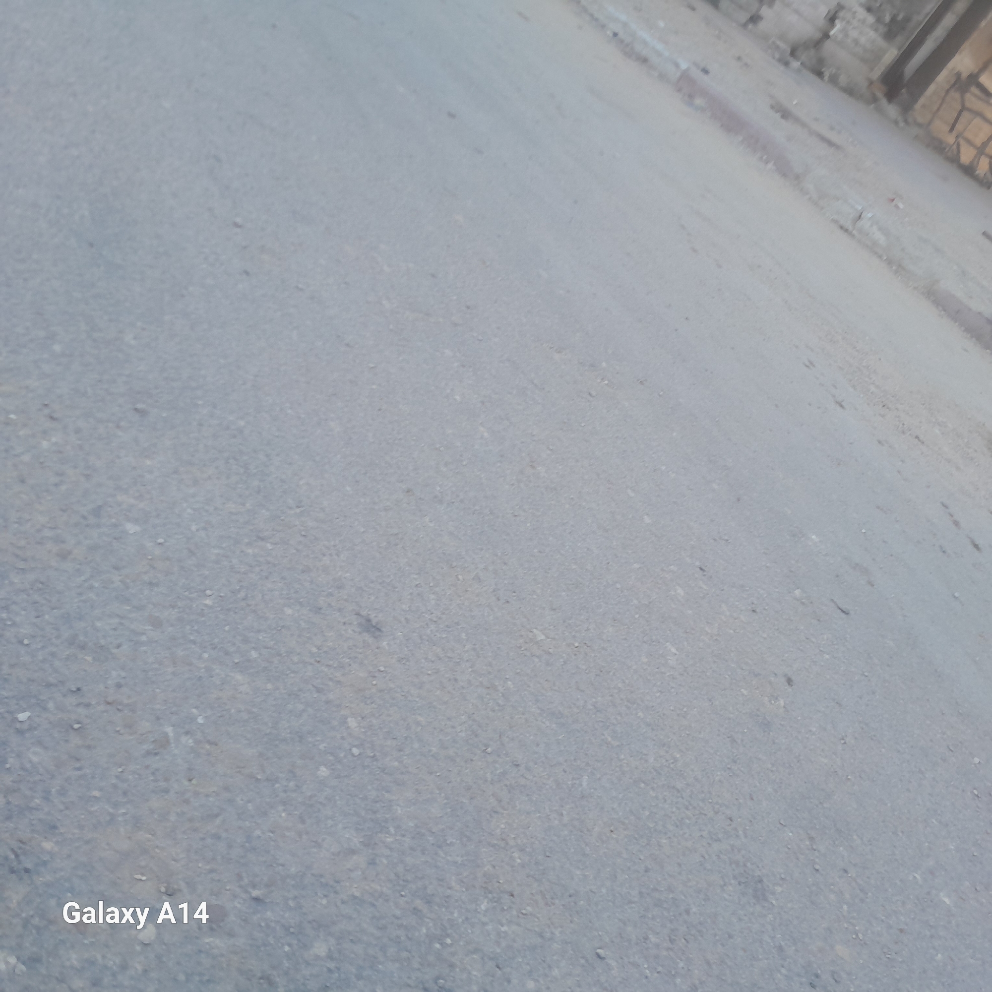 The image displays a tilted view of an asphalt road, likely in Khan Yunis, Palestinian Territories. The road surface is grey and textured, with visible small stones and imperfections. A curb runs along the right side of the road, made of lighter colored material with a reddish-brown hue in places, suggesting wear or material change. In the upper right background, dilapidated structures are visible, appearing to be made of concrete and brick, some with damage. The overall scene is outdoors, under what appears to be natural lighting. The bottom left corner of the image contains the text "Galaxy A14" in white font, indicating the device used to capture the photograph. There are no people or specific actions depicted in the image.