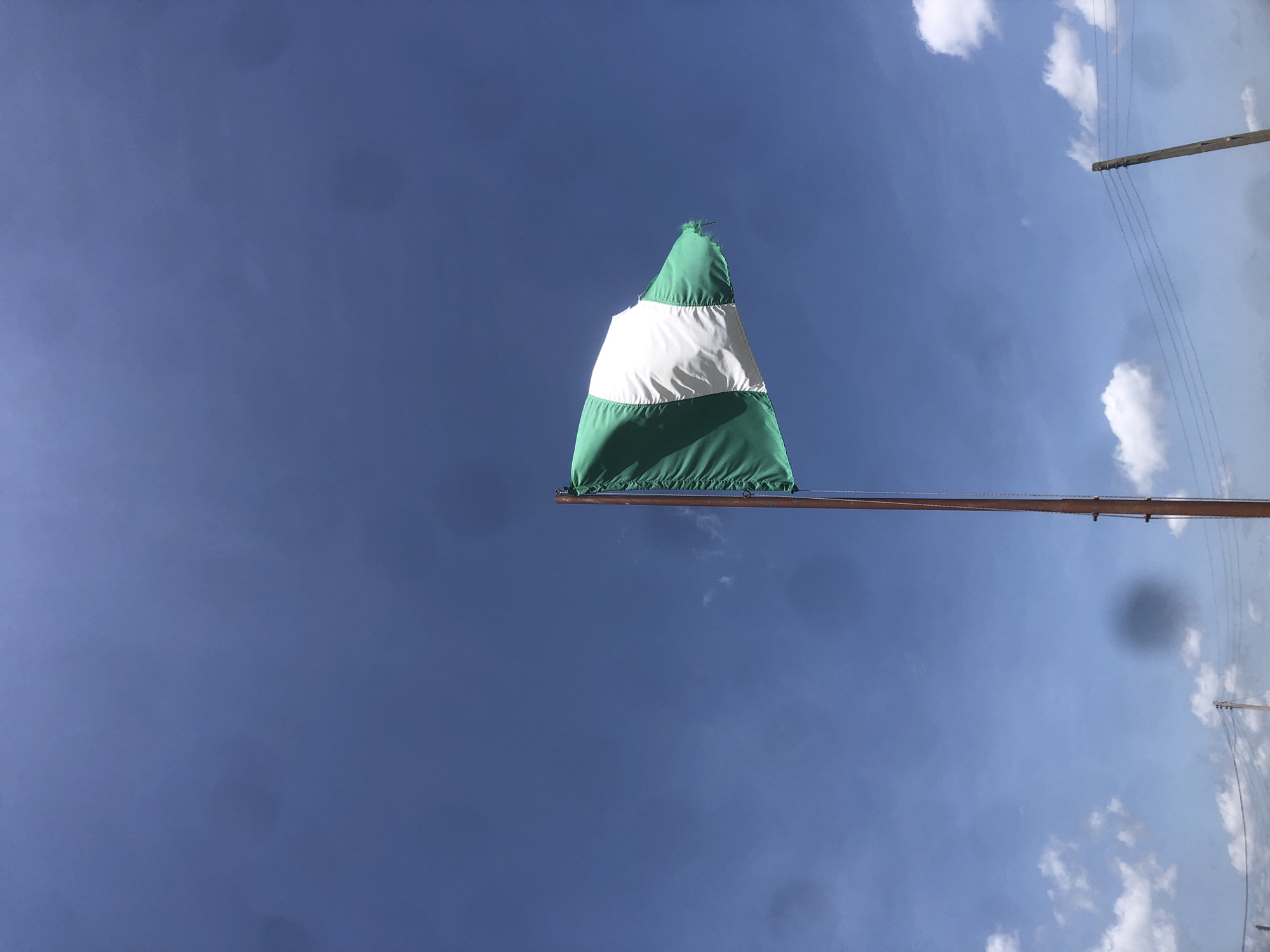 A Nigerian flag, predominantly green and white, flutters against a clear blue sky. The flag is attached to a tall, wooden pole that is visible on the right side of the frame. In the distance, power lines and poles are faintly discernible against a backdrop of scattered white clouds. The setting appears to be an outdoor location in Gwafan, Nigeria.