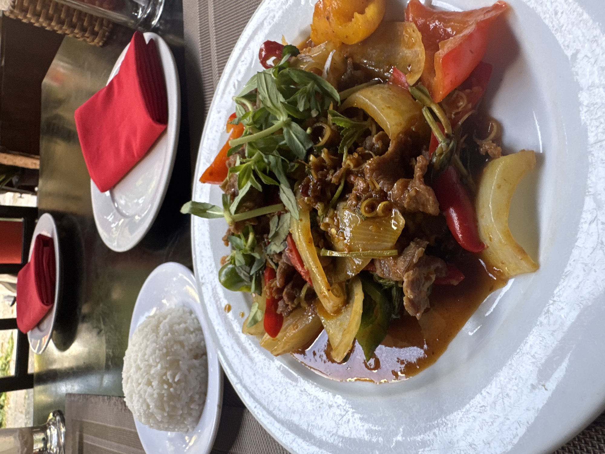 A close-up, slightly overhead shot captures a vibrant meal set on a dark table, likely within a restaurant or dining establishment. The main focus is a large, white circular plate laden with a colorful stir-fry dish. This dish features slices of meat, large pieces of yellow onion, red bell peppers, and red chili peppers, all coated in a rich, dark brown sauce. A generous topping of fresh green herbs, possibly basil, adorns the center of the dish, adding a fresh contrast.
To the left of the main plate, a smaller white plate holds a neatly formed dome of white, fluffy rice. Further back and to the upper left, another white plate holds a bright red, folded cloth napkin. The plates all feature a subtly textured or ridged rim. The table surface is dark, with a visible woven texture on the right side, suggesting a placemat or table runner. Reflections in the background imply shiny surfaces or a window nearby, indicating a well-lit, daytime setting. No people or visible text are present in the image.