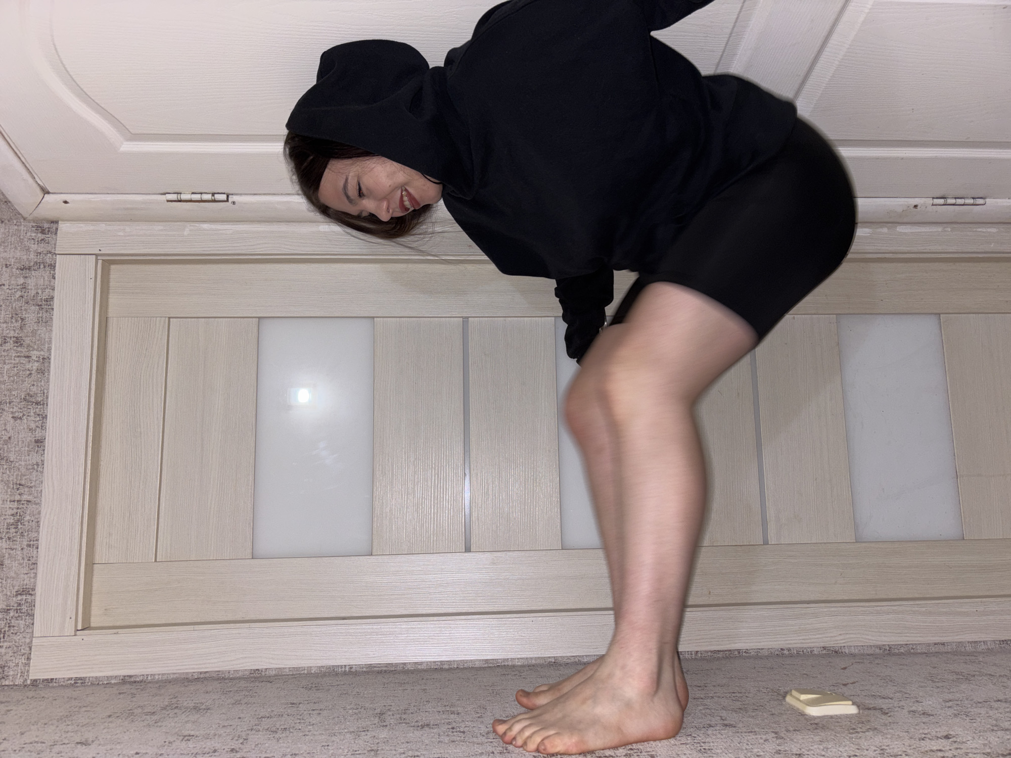 The image, when viewed in its correct upright orientation, captures a young woman bending forward inside what appears to be a room in Perm, Russia. She is wearing a casual outfit consisting of a plain black hooded sweatshirt and black biker shorts. Her face is visible in profile, and she is smiling or laughing as she looks downwards. Her dark hair frames her face. Her bare feet are placed on a light grey or beige carpet that has a subtle, textured pattern.
The background features a light-toned wooden door with multiple vertical panels and a rectangular frosted glass insert on its right side. A bright reflection, likely from an overhead light source, is visible on the glass. The door is framed by a white, slightly decorative architrave or door frame. A silver metallic hinge can be seen attached to this frame near the top left corner of the door.
On the carpet, near the woman's right foot, there is a small, light-colored rectangular object, possibly a bar of soap or a small block. The woman's bent posture combined with her joyful expression suggests a playful moment, perhaps looking at an item on the floor or striking a pose for the camera. The lighting indicates an indoor setting, likely illuminated by artificial light. There is no visible text within the image.