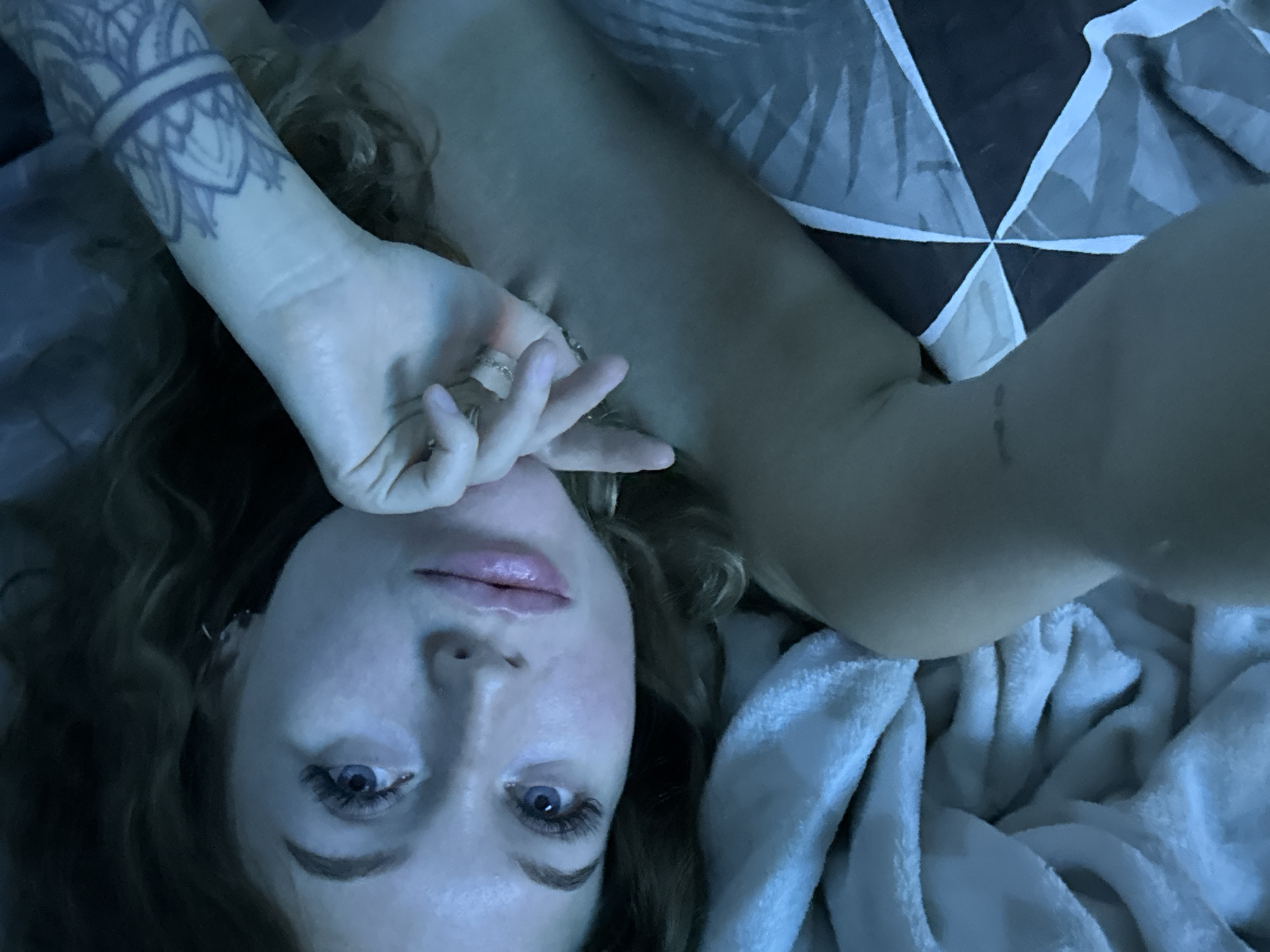 A woman with fair skin, blue eyes, and light brown hair lies on her back, appearing to be on a bed. She is looking directly at the camera with a hand raised near her face. She has several rings on her fingers and a tattoo on her left wrist. The bedding includes a white, fluffy blanket and a patterned sheet with a dark base and geometric white shapes.
There is no indication of the location being Chaikovsky, Russia.