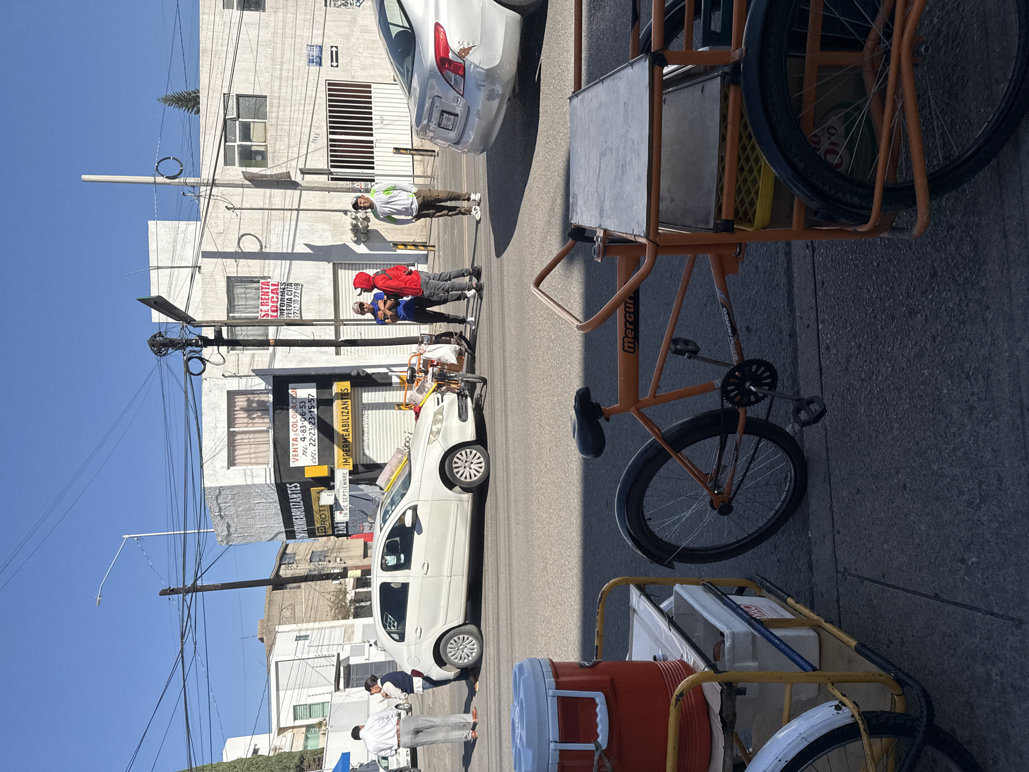 This image captures a bright, sunny street scene in an urban area of Puebla, Mexico. The perspective is tilted, making the street appear as if it runs vertically.
In the foreground and to the right, two pushcarts are visible. One is an orange bicycle-cart with a metal platform and baskets, featuring the brand "Mercur" on its frame. Another cart, partially visible at the bottom, holds a large red and white cooler and a white foam box, suggesting it is used for selling refreshments or food.
Several individuals are present in the midground, standing on the sidewalk and the street. Three men are grouped near a parked white sedan; one wears a red hoodie, another a blue shirt, and the third a white shirt with light pants. Further down the street, another man in a white shirt stands alone. The people appear to be congregating or engaged in casual conversation.
The background consists of multi-story buildings lining the street, characterized by light-colored facades, windows, and roll-down security gates. Numerous power lines crisscross the clear blue sky, connecting to a utility pole. The bright sunlight casts strong shadows, indicating it is likely mid-day.
Visible text on signs advertises local services and properties. A red and white sign reads "SE RENTA LOCAL INFORMES PREVIA CITA 222176 22 69" (Local for Rent, inquiries by appointment). Yellow and black signs promote "VENTA COLOCACION IMPERMEABILIZANTES TEL. 4-83-06-53 CEL. 22-23-23-15-57" (Sale and Installation of Waterproofing) and "16 DE SEPTIEMBRE IMPERMEABILIZANTES." Partial text "PROTEA IM" is also visible. The overall scene suggests a typical bustling street with commercial activity.
