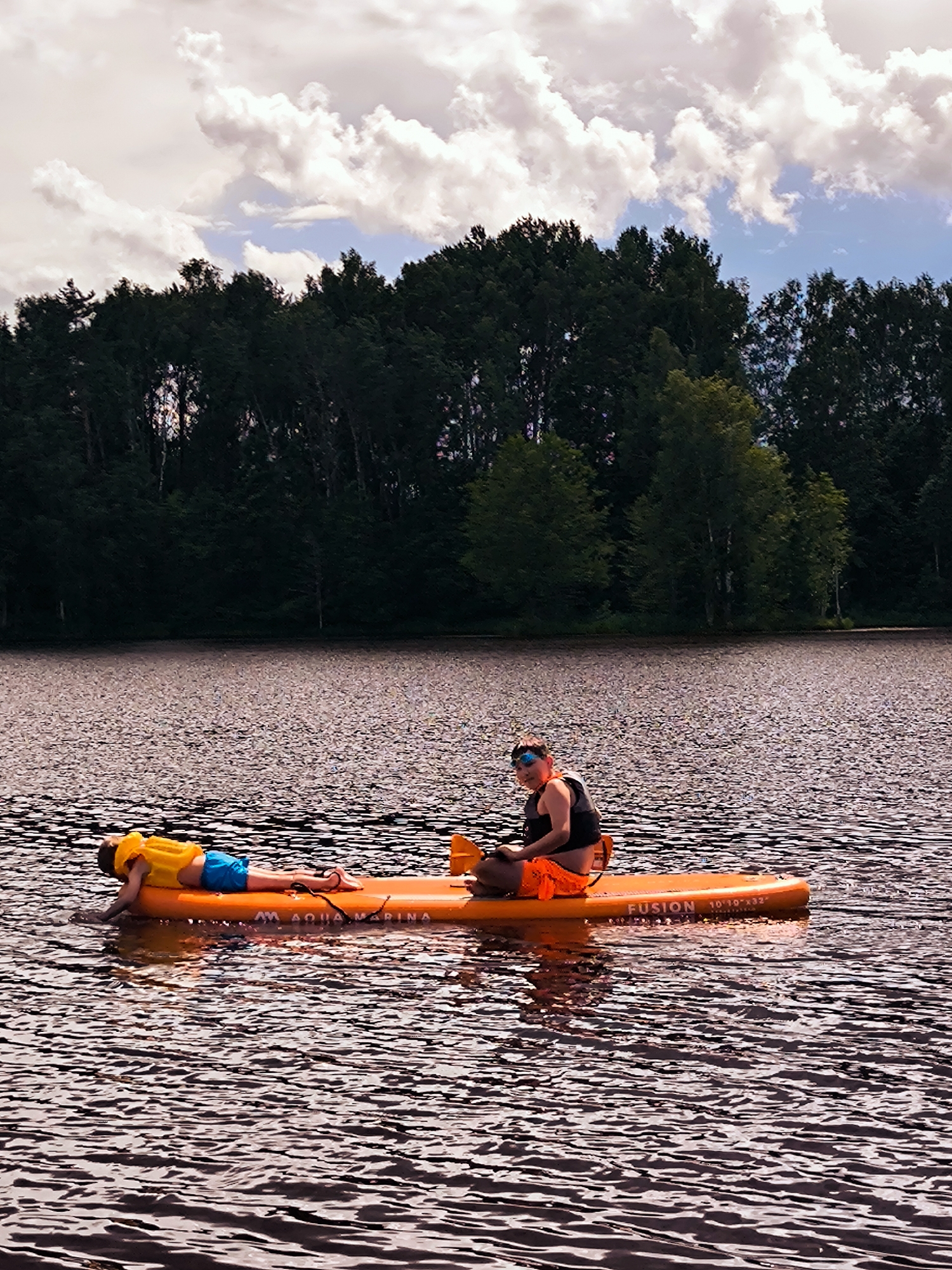 Two people are on an orange paddleboard on a body of water, likely a lake, with a line of dark green trees along the far bank under a sky with scattered white clouds. In the foreground, the water has a rippled surface with dark reflections. On the paddleboard, a person wearing a yellow life vest and blue shorts is lying face down. To their right, a person wearing a black top, orange shorts, and sunglasses is sitting and holding a paddle. The paddleboard has "AQUA MARINA" and "FUSION 10'10"x32"" printed on it. This scene is consistent with being at the Daugava River near Vecumnieki, Latvia, a popular spot for watersports.