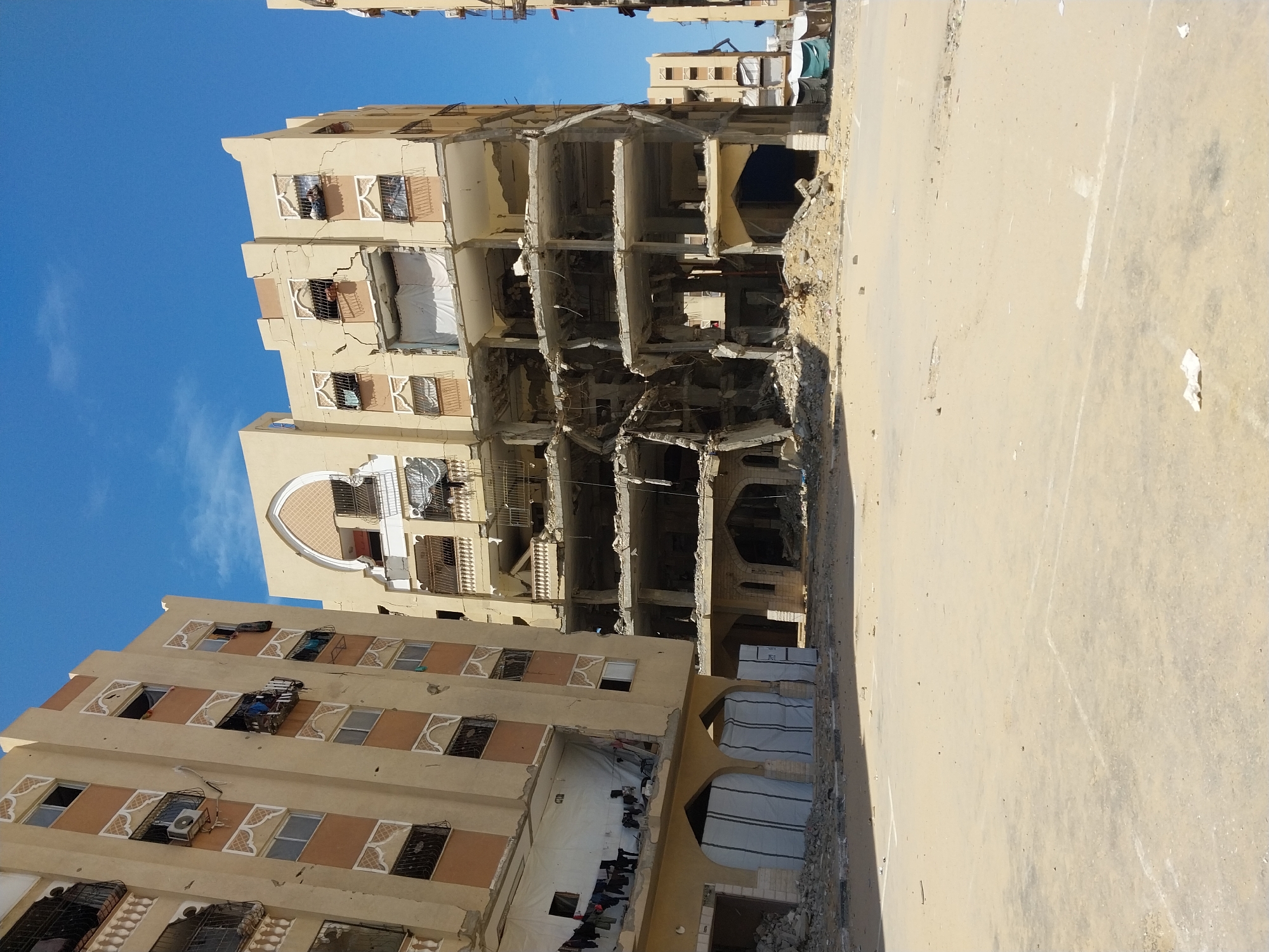 The image depicts a devastated urban landscape in Khan Yunis, Palestinian Territory, characterized by heavily damaged multi-story residential buildings under a clear blue sky. In the foreground, a wide, flat, sandy, and dusty area, possibly a street or plaza, shows faint white markings and scattered debris.
The central and most prominent structure is a multi-story building with its central section collapsed inward, leaving exposed, fractured concrete slabs and twisted rebar across multiple levels. The upper floors of this building display severe structural cracks, missing window panes, and damaged balconies, some fitted with security grates.
To the left, another multi-story building, constructed of light-colored material, exhibits extensive surface damage consistent with impact, including numerous small pockmarks on its facade. Its windows are either broken or missing, and some balconies are damaged. On a lower floor, a large opening is partially covered by white sheeting, with laundry hung to dry on a line. The ground level features arched entryways, some of which are covered with white tarps, with rubble accumulated at the base.
A third building, partially visible on the far right, shows similar damage and construction style. Piles of concrete rubble are scattered at the base of all visible structures. No individuals or active vehicles are present in the scene.