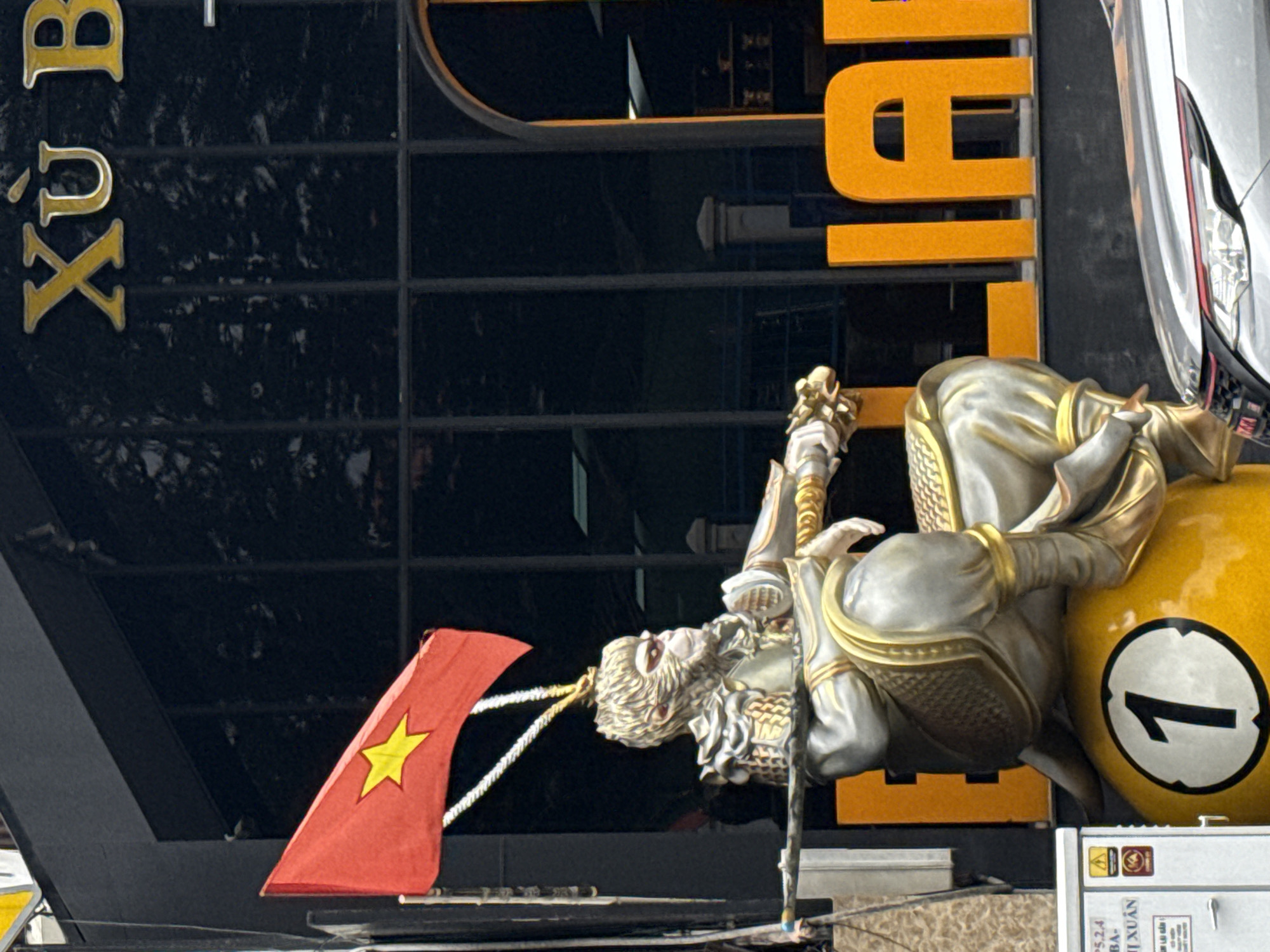 A statue of Sun Wukong, also known as the Monkey King, sits on top of a giant pool ball with the number 1 on it. Sun Wukong is depicted wearing ornate armor with gold accents, holding a staff. A Vietnamese flag flies above the statue. Behind the statue, there is a dark building with large windows and a sign that reads "XÙ B" in gold letters. To the right of the statue, large orange letters spell out "LI". The scene appears to be a street in Da Lat, Vietnam, indicated by the presence of the Vietnamese flag and the style of the building and signage. A silver car is parked in the foreground.