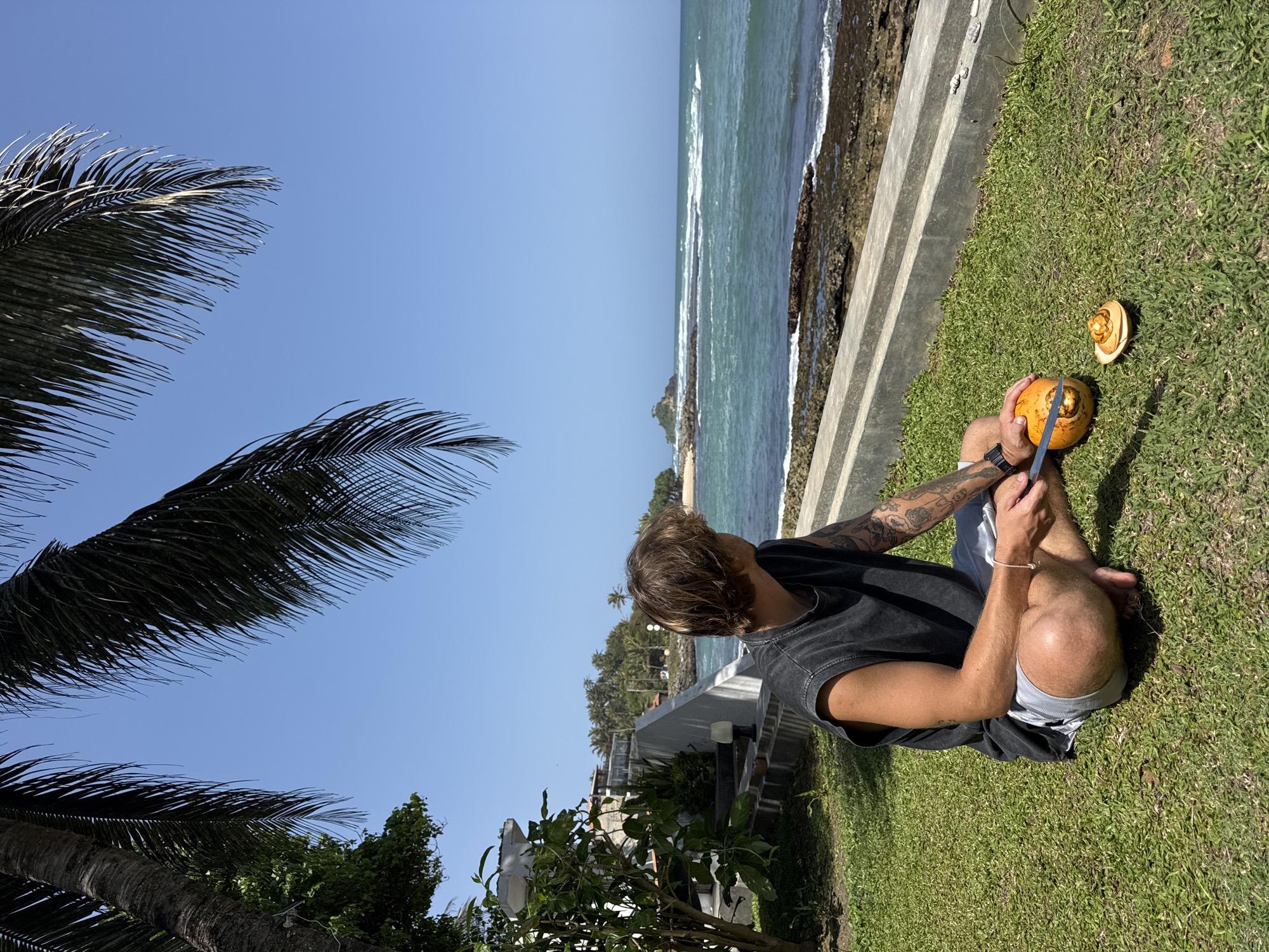 A person is sitting on a grassy lawn in front of a concrete barrier. The person is wearing a dark sleeveless t-shirt and light-colored shorts. They are holding a knife and cutting into a yellow coconut. A portion of the coconut has been removed and is placed next to it. The person has tattoos on their arm. In the background, there is the ocean with waves crashing on the shore and a rocky coastline. There are palm trees with fronds extending into the frame from the top. A building with stairs is visible to the left of the person. The sky is clear and blue. The location is the city of Goviyapana, Sri Lanka.