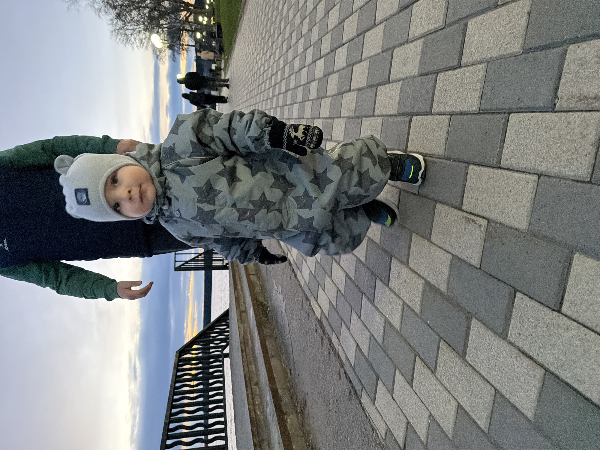 A low-angle, portrait-oriented photograph, rotated 90 degrees clockwise, captures a young child standing on a paved walkway in Votkinsk, Russia, during dusk or early evening. The child, likely a toddler, is at the center, looking directly at the camera with a neutral expression. They are dressed warmly in a light grey hat with ear-like flaps, a matching full-body snowsuit or overall adorned with dark grey star patterns, dark patterned mittens, and dark sneakers with bright green/yellow accents.
An adult's arm and torso, wearing a green long-sleeved top under a dark jacket, are visible on the left, close to the child, suggesting they are accompanying the child. A partially visible brand name "joep" can be seen on the adult's green sleeve.
The setting is an outdoor promenade or boardwalk. The ground is paved with alternating light and dark grey rectangular blocks. To the left, a dark metal railing with wavy horizontal bars separates the walkway from a body of water, likely a lake or river, which reflects the pale, muted colors of the sky near the horizon. The sky transitions from light grey overhead to soft blues and faint yellows/oranges closer to the horizon, indicating either sunset or just after.
In the background, bare trees and illuminated streetlights are visible, along with silhouettes of other people further down the path, suggesting a public space. The overall atmosphere is cool and peaceful.