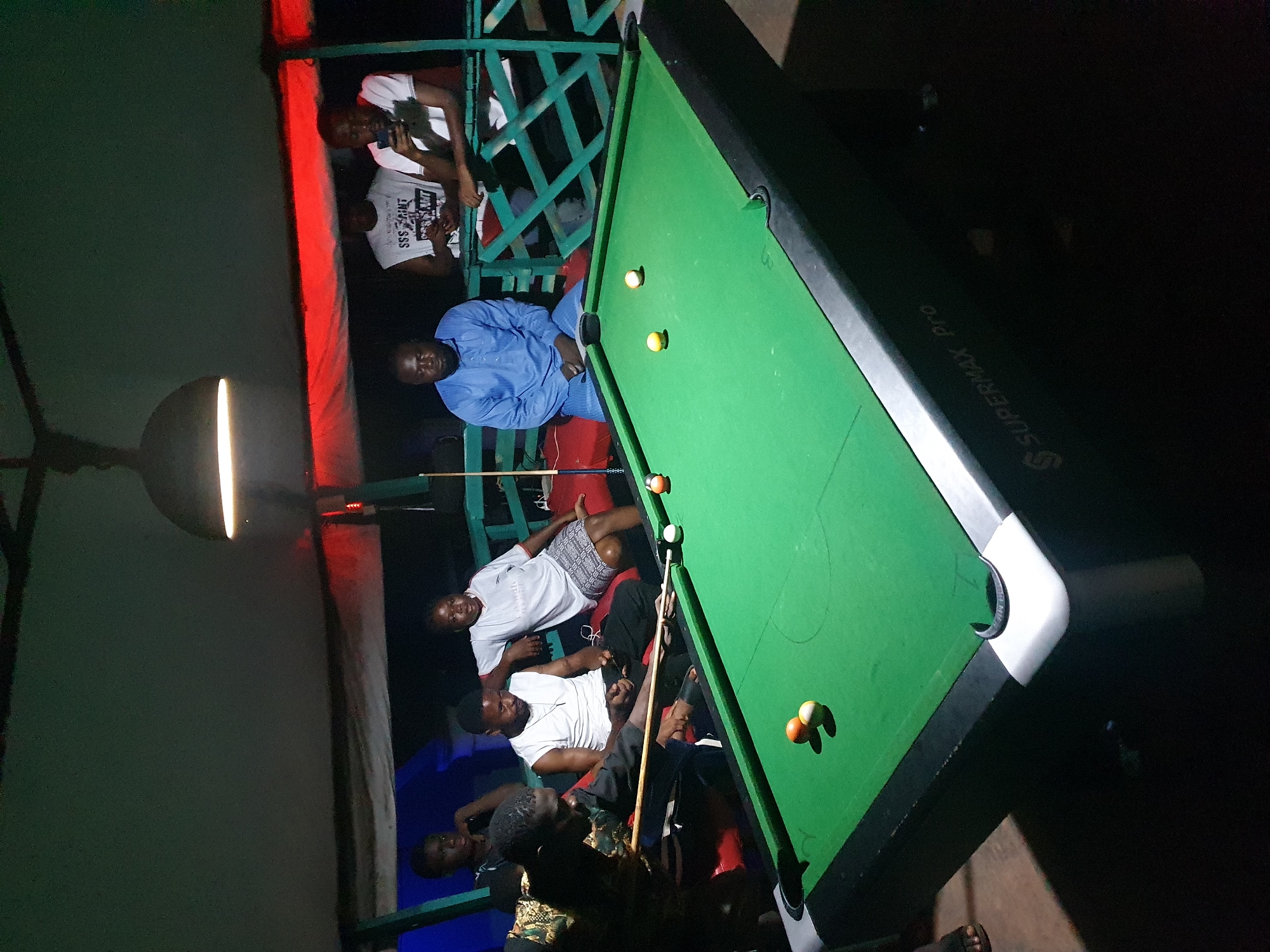 The image depicts a group of men gathered around a pool table in Lokoja, Nigeria. A lit pool table dominates the center of the frame, with several billiard balls scattered across its green surface. Four men are seated or reclining along one side of the table, while three others stand at the opposite side. One of the standing men holds a pool cue. A light fixture is suspended above the pool table. A wooden fence and portions of a structure painted in red and blue form the background. The overall scene suggests an evening gathering at an outdoor recreational space. The pool table is a Supermax Pro.