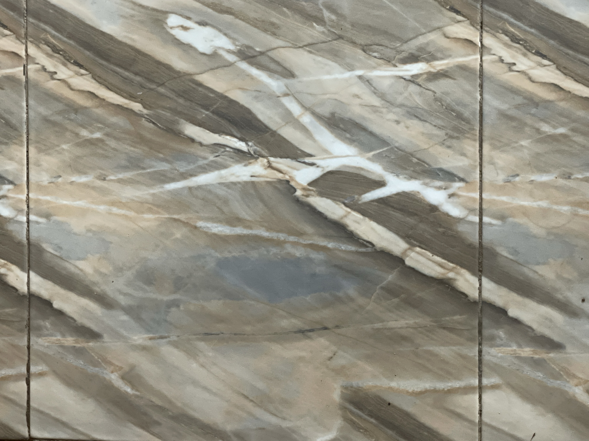 The image shows a close-up of two ceramic tiles with a marbled pattern. The tiles are beige, brown, and gray with white veining, mimicking the appearance of natural stone. A thin, dark grout line separates the two tiles horizontally across the center of the image. The lighting is even, with no harsh shadows. The surface appears to be clean and smooth. This image does not contain any information or context related to Kakau, Nigeria.