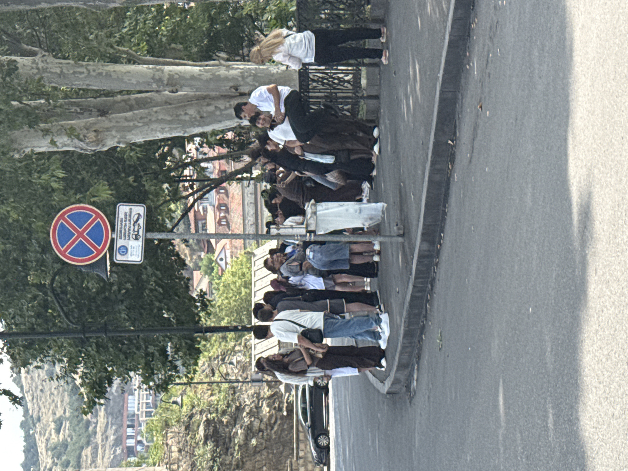 A group of young people are standing and sitting on a sidewalk next to a road in Tbilisi, Georgia. A black car is parked on the side of the road. A traffic sign with a red circle and a blue cross is visible above the group. The background shows buildings on a hillside. The weather appears to be warm and sunny.