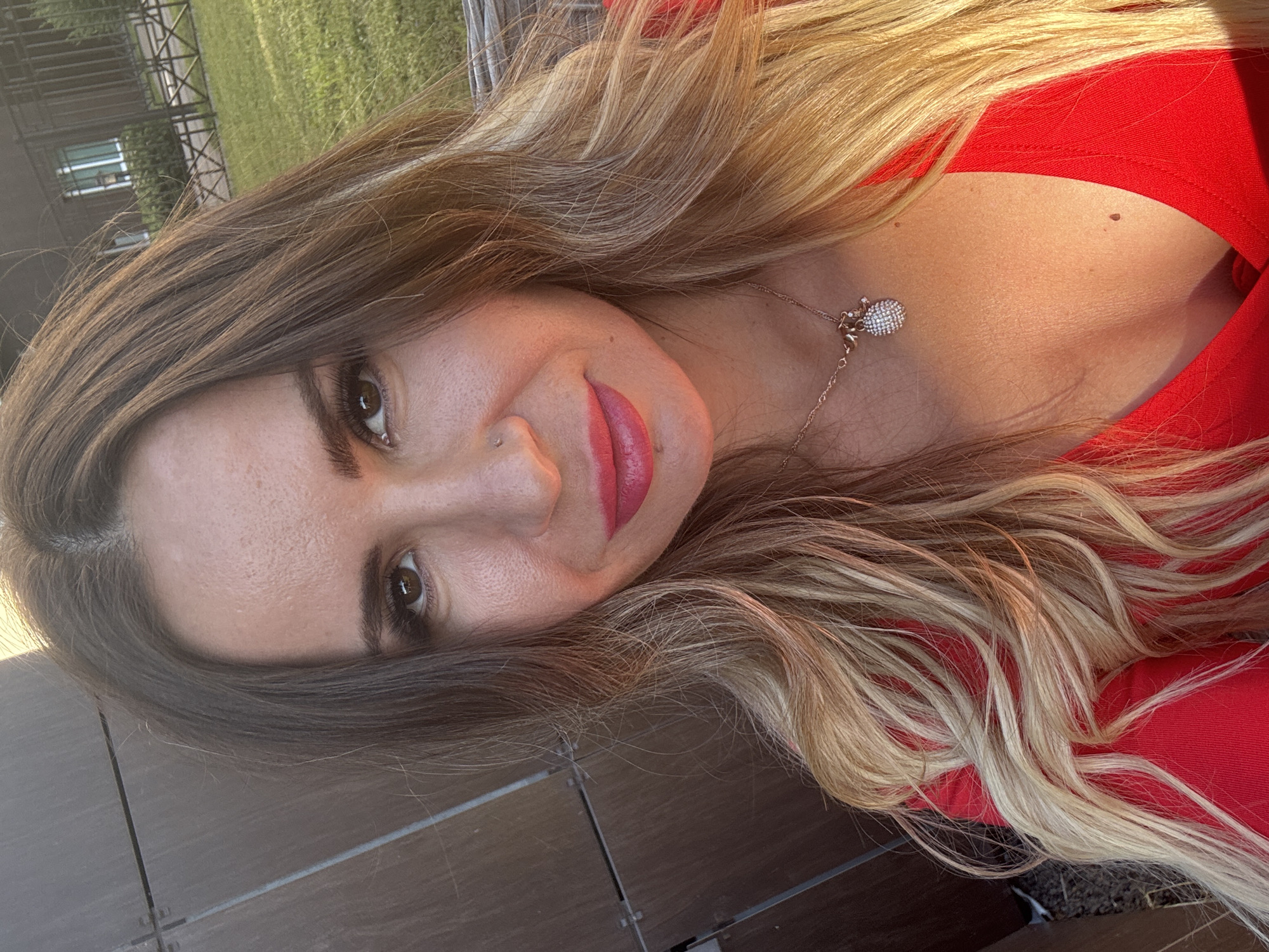 This media file features a close-up, rotated portrait of a woman outdoors. The woman has long, layered blonde and brown hair that frames her face. She is wearing a red top and a delicate necklace with a sparkling charm. Her makeup includes pink lipstick and mascara, and she has a nose piercing. The background is blurred, but suggests an outdoor setting with greenery and a building visible in the distance. The lighting indicates it is daytime, likely sunny, casting warm tones on her skin. The overall mood appears relaxed and casual. There is no visible text in the image.