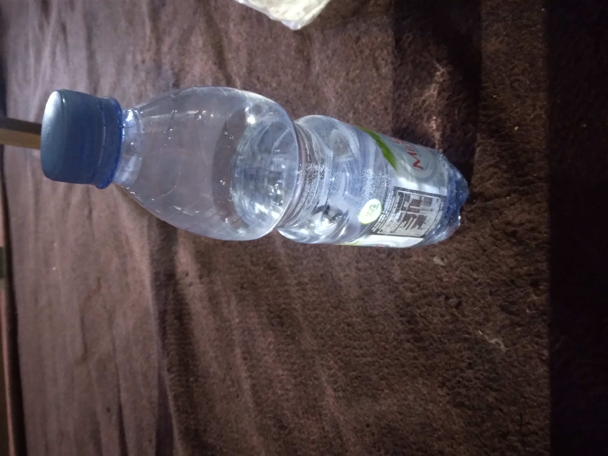 A clear plastic bottle of mineral water with a blue cap stands upright on a textured brown surface. The bottle is partially filled with water, and tiny bubbles are visible. A label with text and green accents is affixed to the lower portion of the bottle. The background is a dark, textured surface, suggesting it might be a blanket or rug. The lighting is dim, casting some shadows. The image appears to have been taken in an indoor setting, possibly a home or restaurant, in Pacet, Indonesia.