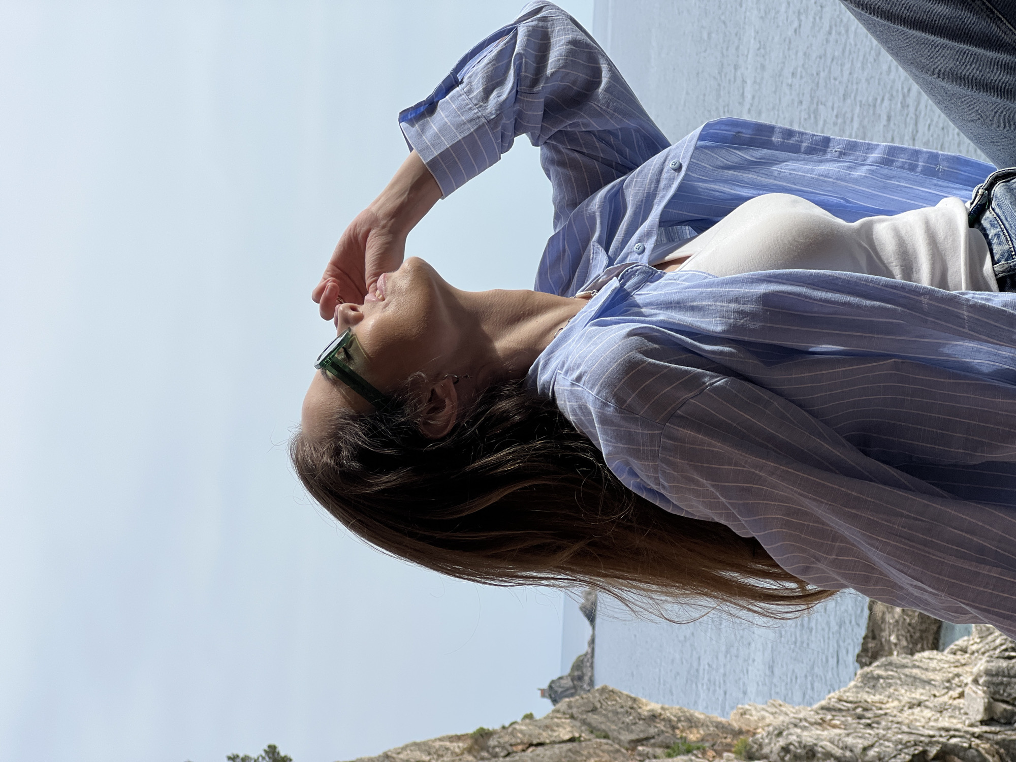 A woman is posing outdoors, possibly on a cliff or high point in Perazića Do, Montenegro. She is wearing a striped light blue shirt over a white top and jeans, and green-rimmed glasses. Her left hand is raised near her face. In the background is a blue sky, the ocean, and rocky terrain.