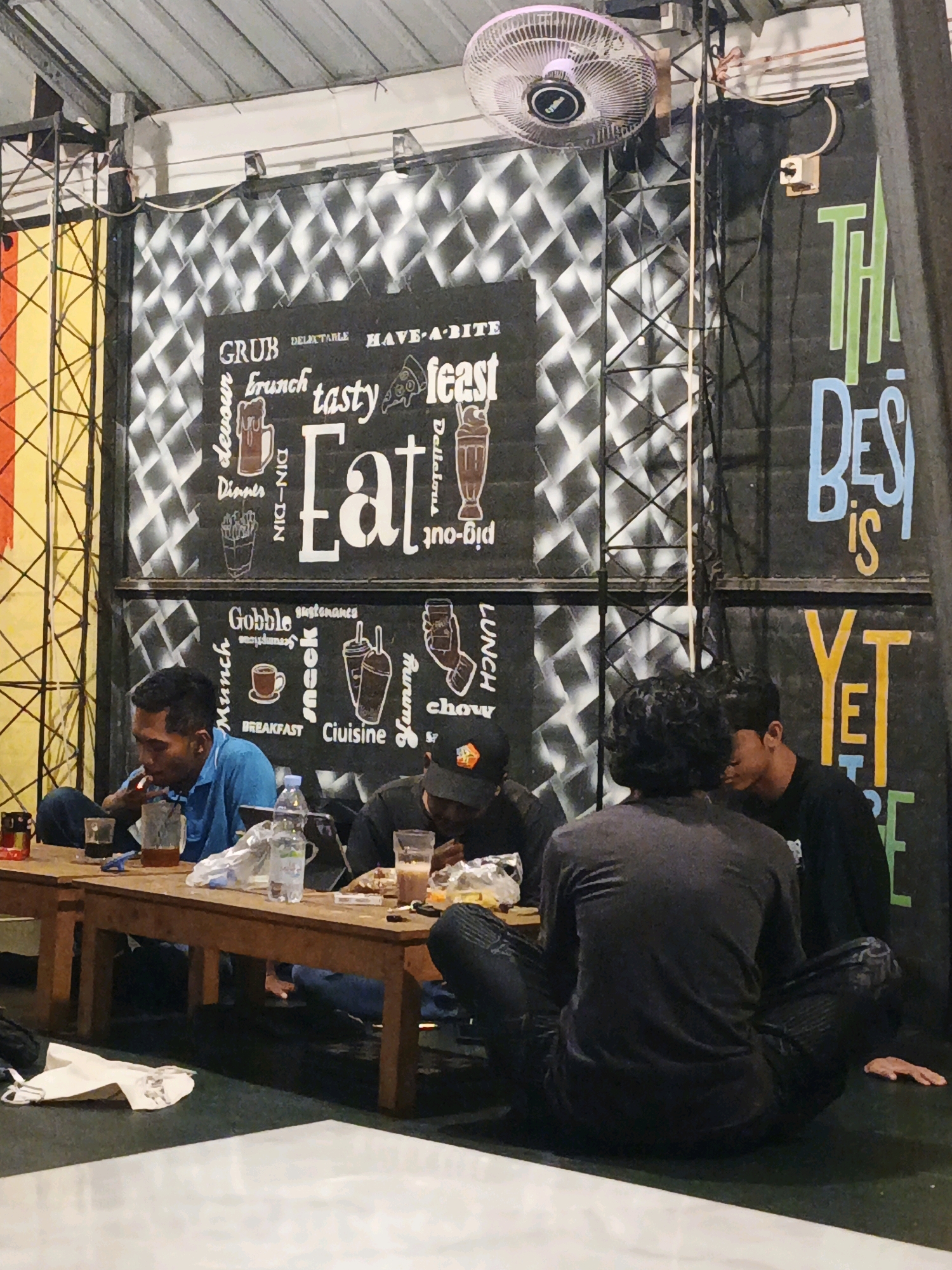In Ketegan, Indonesia, a dimly lit indoor dining area features a long wooden table where four young men are seated. Three of the men are visible in the foreground and midground, engaged in eating or drinking. The man in the bright blue shirt is smoking. The wall behind them is decorated with a patterned mural and stylized lettering that includes "GRUB", "delicious", "brunch", "tasty", "feast", "Eat", "Gobble", "Munch", "BREAKFAST", "Ciuisine", "Yummy", and "CHOW". The right side of the wall has larger, colorful lettering that reads "THE BEST IS YET TO COME". A ceiling fan is visible overhead. The overall atmosphere appears casual and relaxed, characteristic of a local eatery.