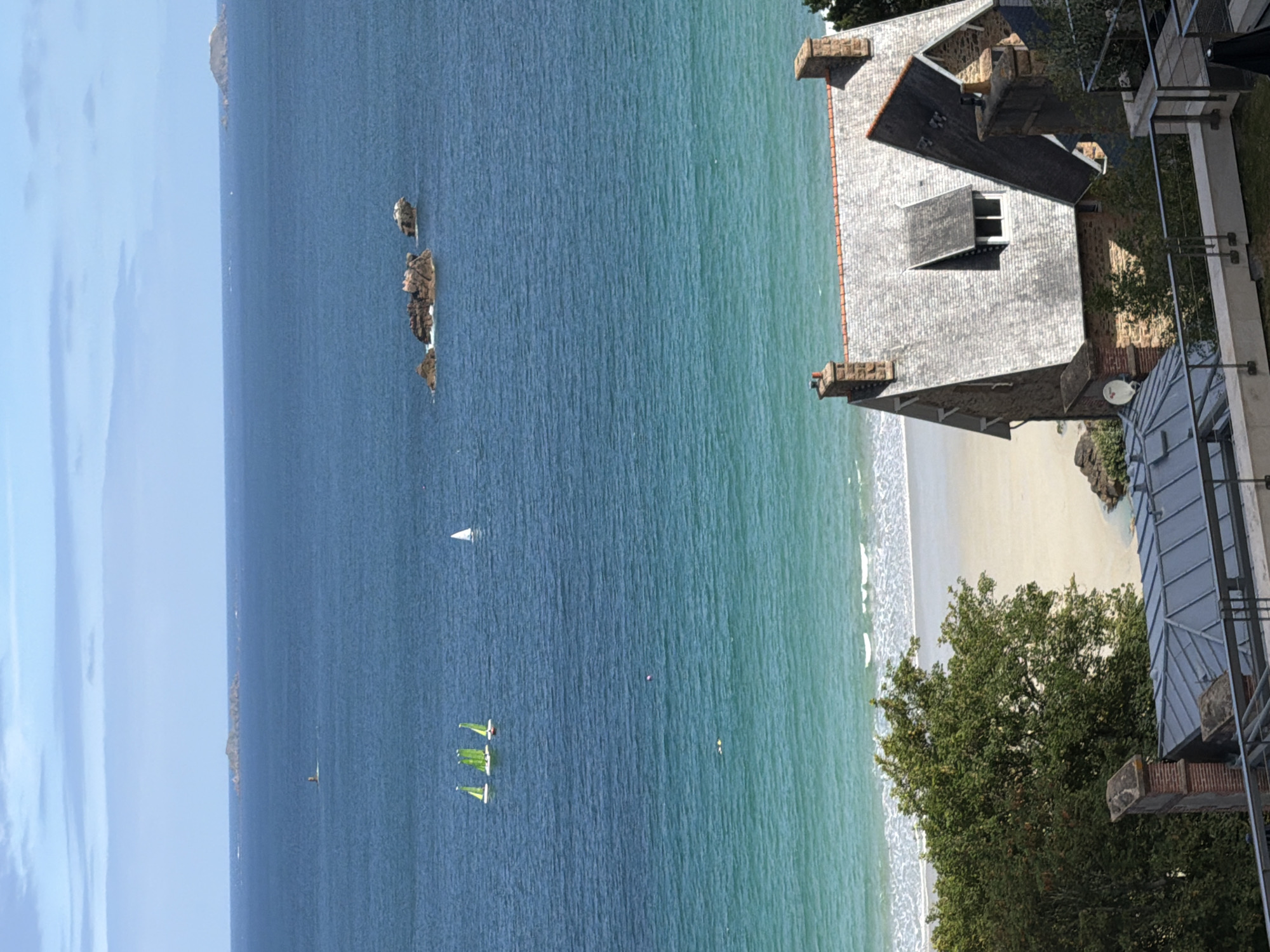 The image displays a picturesque coastal scene in La Clarté, France. The foreground features a stone house with a grey slate roof and a terra cotta chimney. A metal railing and a section of a modern balcony or extension are visible to the right of the house. Lush green trees grow on the grounds surrounding the house.
Beyond the house, the turquoise waters of the ocean meet a sandy beach, where gentle waves break. Several small sailboats with green sails are navigating the water. In the distance, a cluster of rocks emerges from the sea, and further out, a hazy outline of land can be seen. The sky is a pale blue with hints of white clouds. The lighting suggests it is daytime, with clear, pleasant weather. No people are explicitly visible, but the presence of the sailboats implies recreational activity on the water. The overall impression is one of a tranquil, scenic coastal setting.