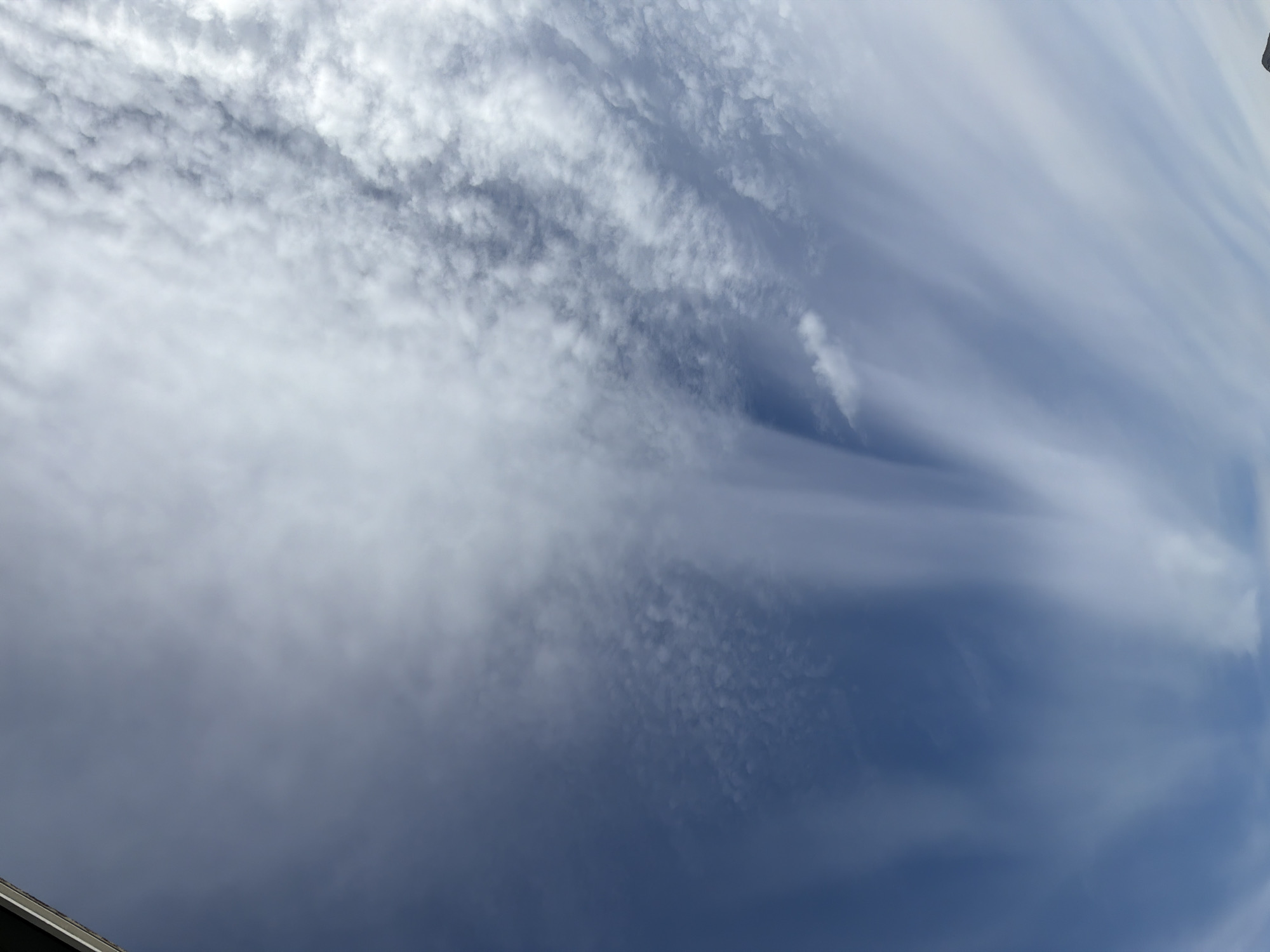 The image shows a sky filled with wispy cirrus clouds and some cumulus clouds. The blue sky is visible in between the clouds. The clouds are thin and spread out, creating a feathery appearance. The lighting suggests it is daytime, likely late morning or early afternoon, with the sun positioned somewhere above and to the right of the frame, casting light on the clouds. The image is oriented vertically, with the bottom of the frame tilted upwards towards the sky. There is a sliver of a dark, angular structure visible along the very top left edge of the image, which could be part of a building or a tall object. No people or specific objects are clearly discernible within the cloud formations or the sky. The scene is consistent with an outdoor environment in Storsteinnes, Norway, on a day with variable cloud cover.